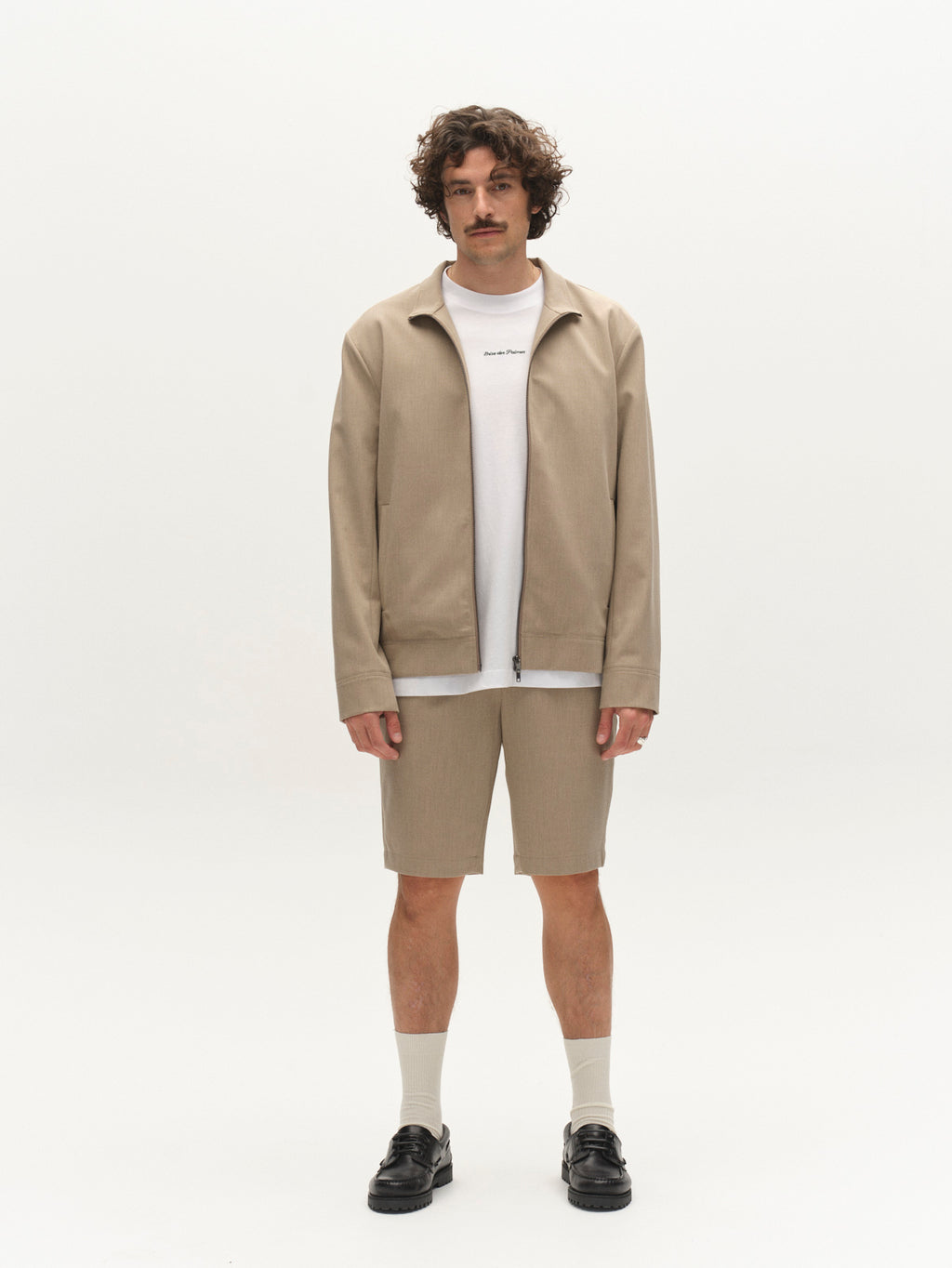 A man with curly hair and a mustache stands against a white background wearing Gabba GABjoli Pas Phin Shorts GRS in Sand Plaza, a beige jacket, white t-shirt, white socks, and black shoes, looking directly at the camera.