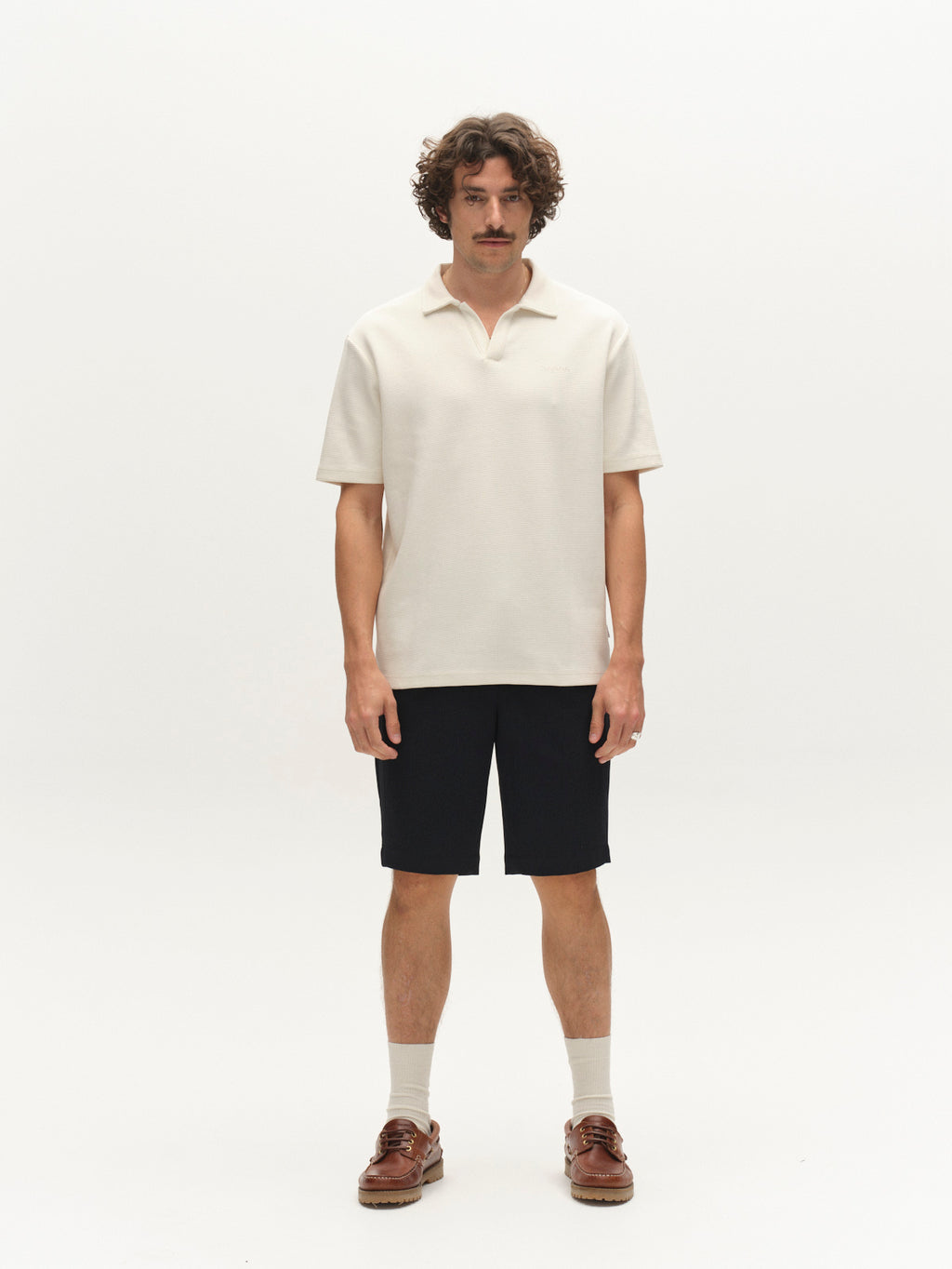A man with curly brown hair and a mustache stands against a white background, wearing a cream short-sleeve polo shirt, Gabba GABjoli Pas Phin Shorts GRS in Blue Black, beige socks, and brown boat shoes.