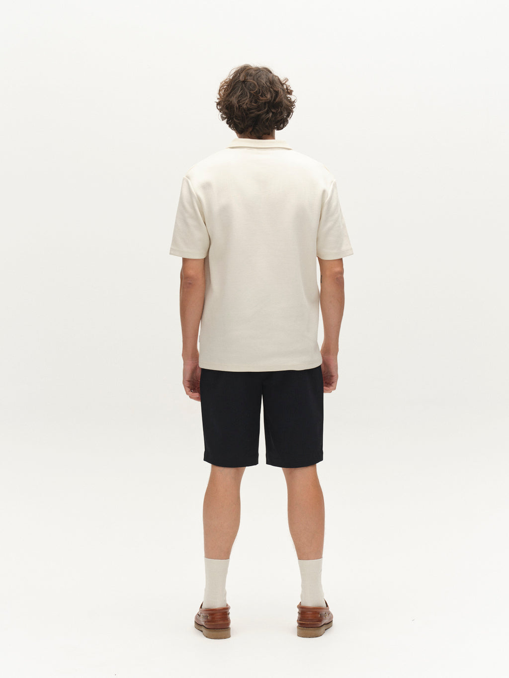 A person with curly hair, facing away, wears a cream short-sleeve polo, Gabba GABjoli Pas Phin Shorts GRS - Blue Black, light socks, and brown shoes against a plain white background.