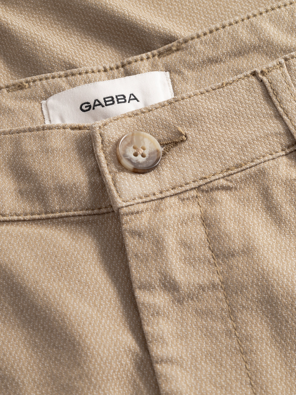 Close-up of GABjoli Nopleats Dale Shorts in sand by Gabba featuring a white Gabba label, light button, and detailed stitching on the waistband and fly area.