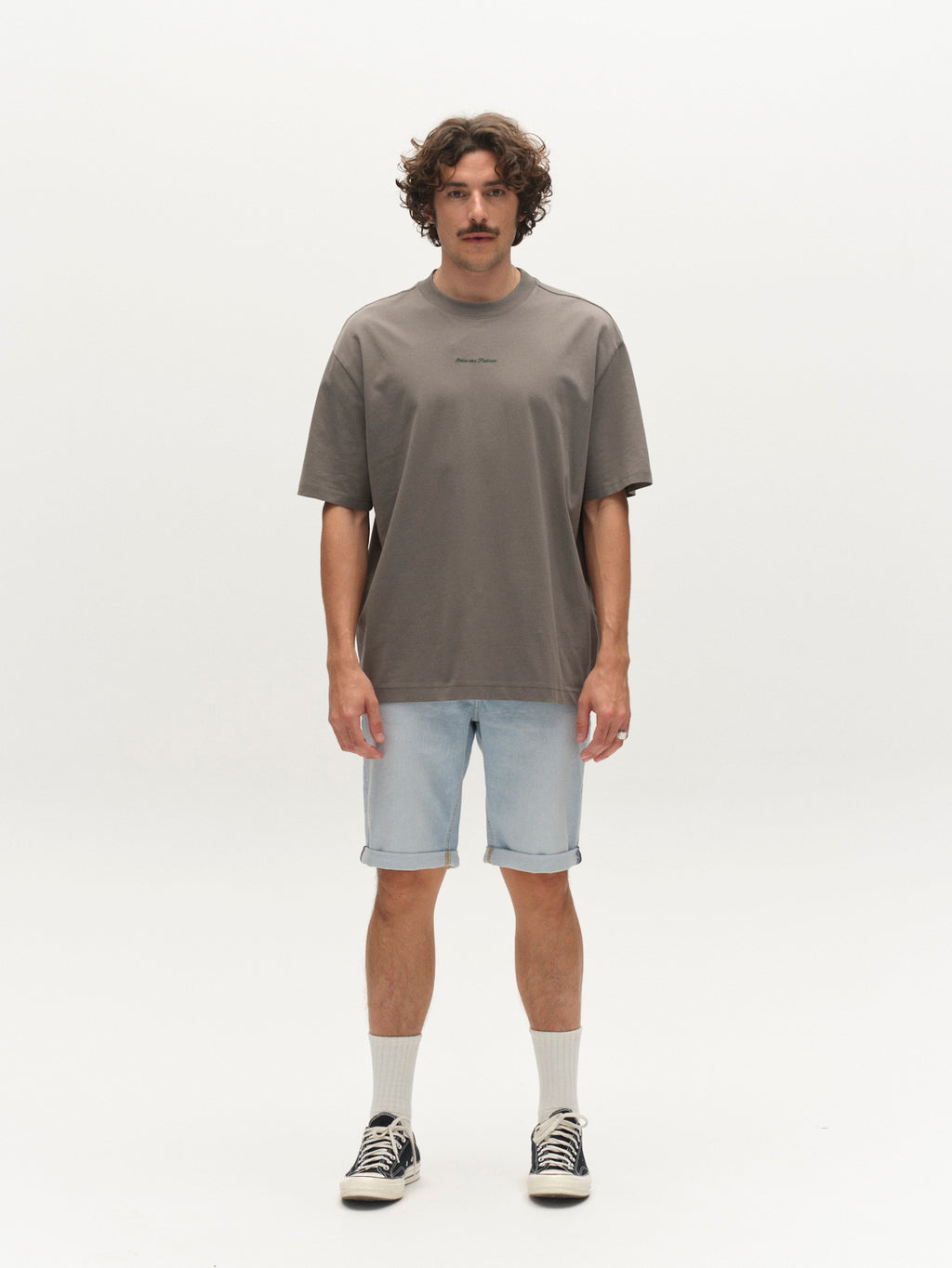 A man with curly hair and a mustache stands against a white background, wearing casual summer style: a loose gray tee, Gabba GABmarkus Derby Shorts in Lt. Blue Denim, white crew socks, and black sneakers.