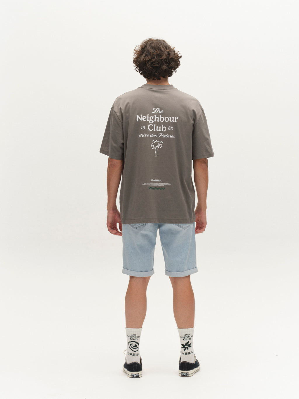 A person with short curly hair faces away, wearing a gray tee, Gabba GABmarkus Derby Shorts in light blue denim, patterned socks, and black shoes—an effortless casual summer look against a plain white background.