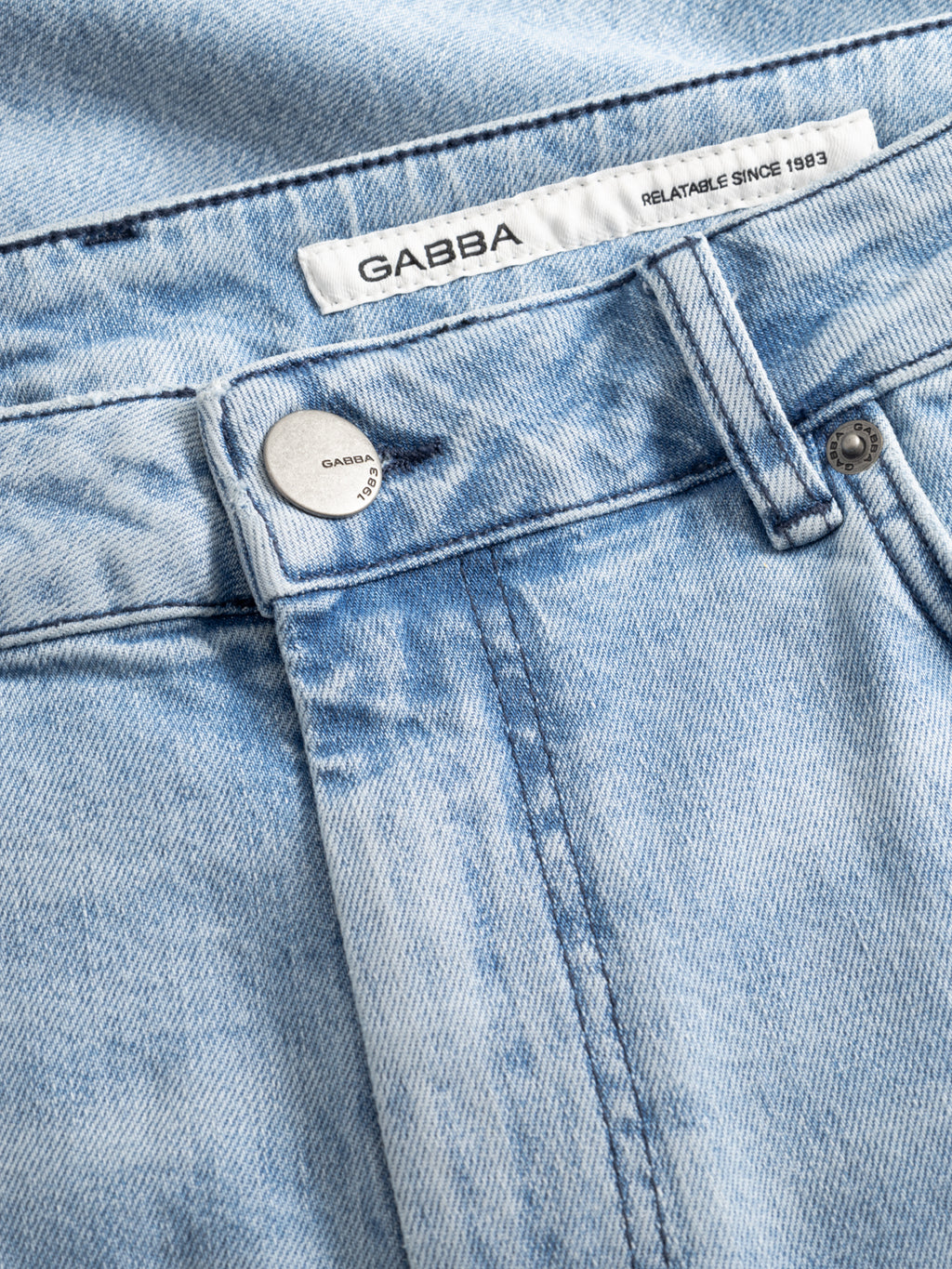 Close-up of Gabba GABmarkus Derby Shorts in mid-blue denim, featuring a metal button with Gabba engraving, belt loops, detailed pocket stitching, and a white label reading 