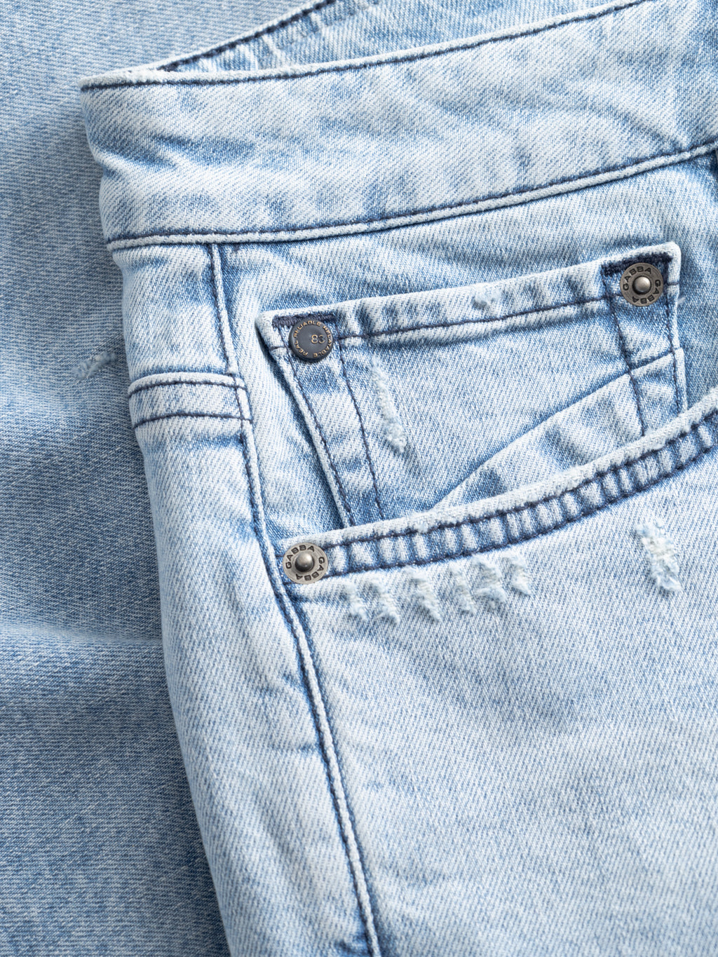 Close-up of Gabba's GABmarkus Derby Shorts in mid blue denim, featuring a front pocket, metal rivets, and subtle distressed accents, laid flat on matching denim.