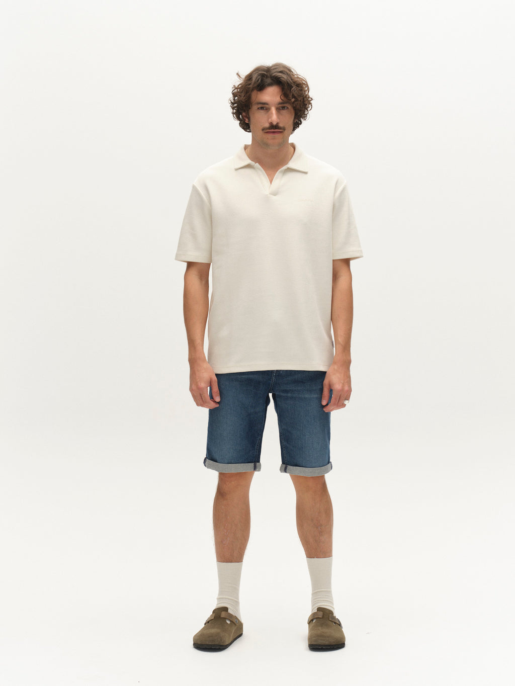 A man with curly hair and a mustache stands facing forward, wearing a cream short-sleeve polo shirt, Gabba GABmarkus Derby Shorts in denim wash, beige socks, and brown sandals against a plain white background.