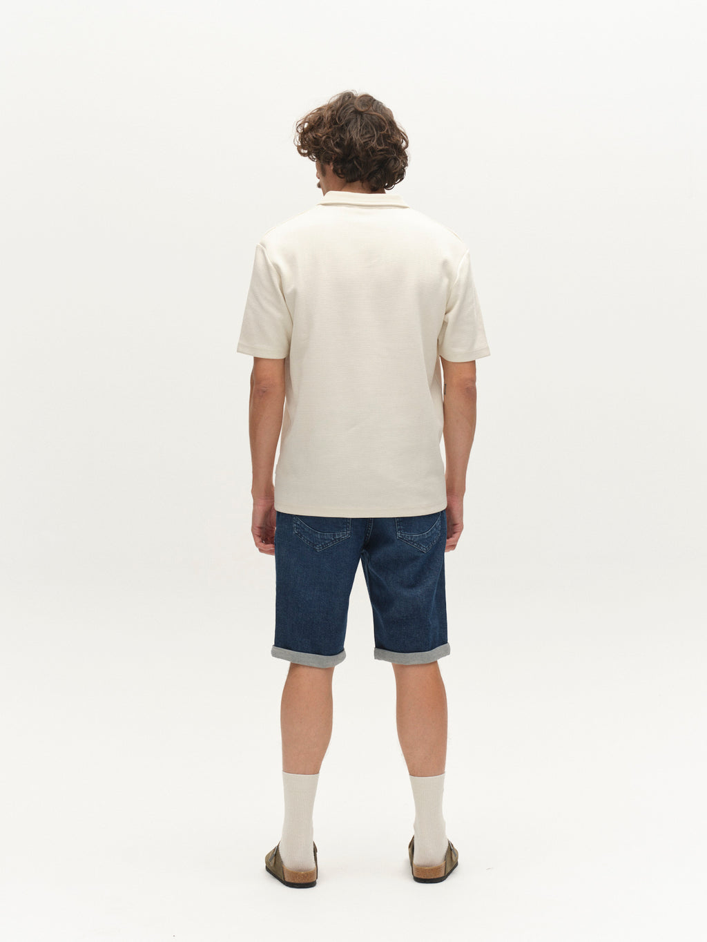 A person with curly hair, facing away from the camera, wears a white short-sleeve shirt and Gabba GABmarkus Derby Shorts in denim wash, plus beige socks and brown sandals, standing against a plain white background.