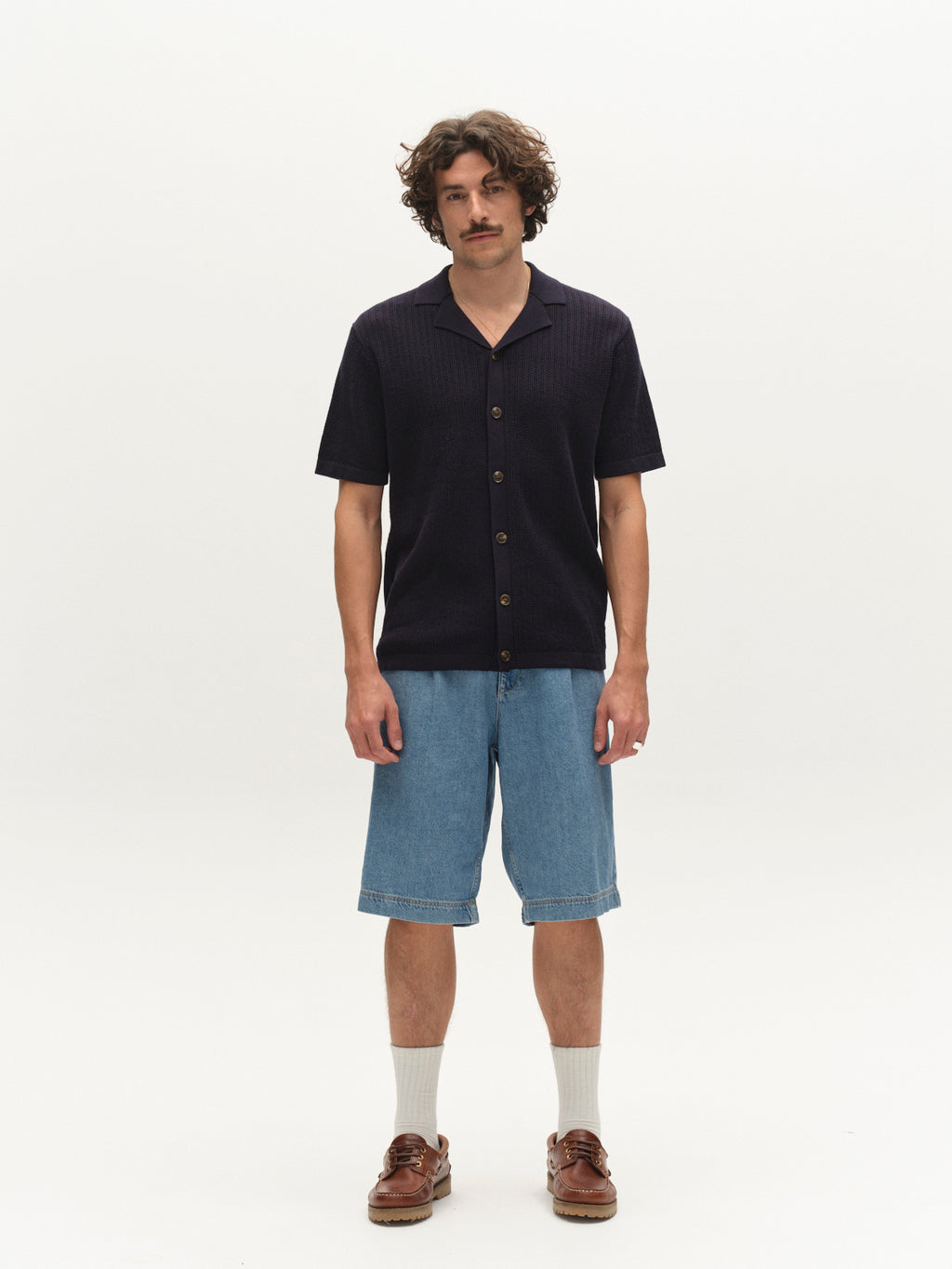 A man with curly hair and a mustache stands against a white background, wearing a dark short-sleeve button-up shirt, Gabba GABmoro PasBrook Shorts in denim wash, white socks, and brown boat shoes. A true summer essential look.