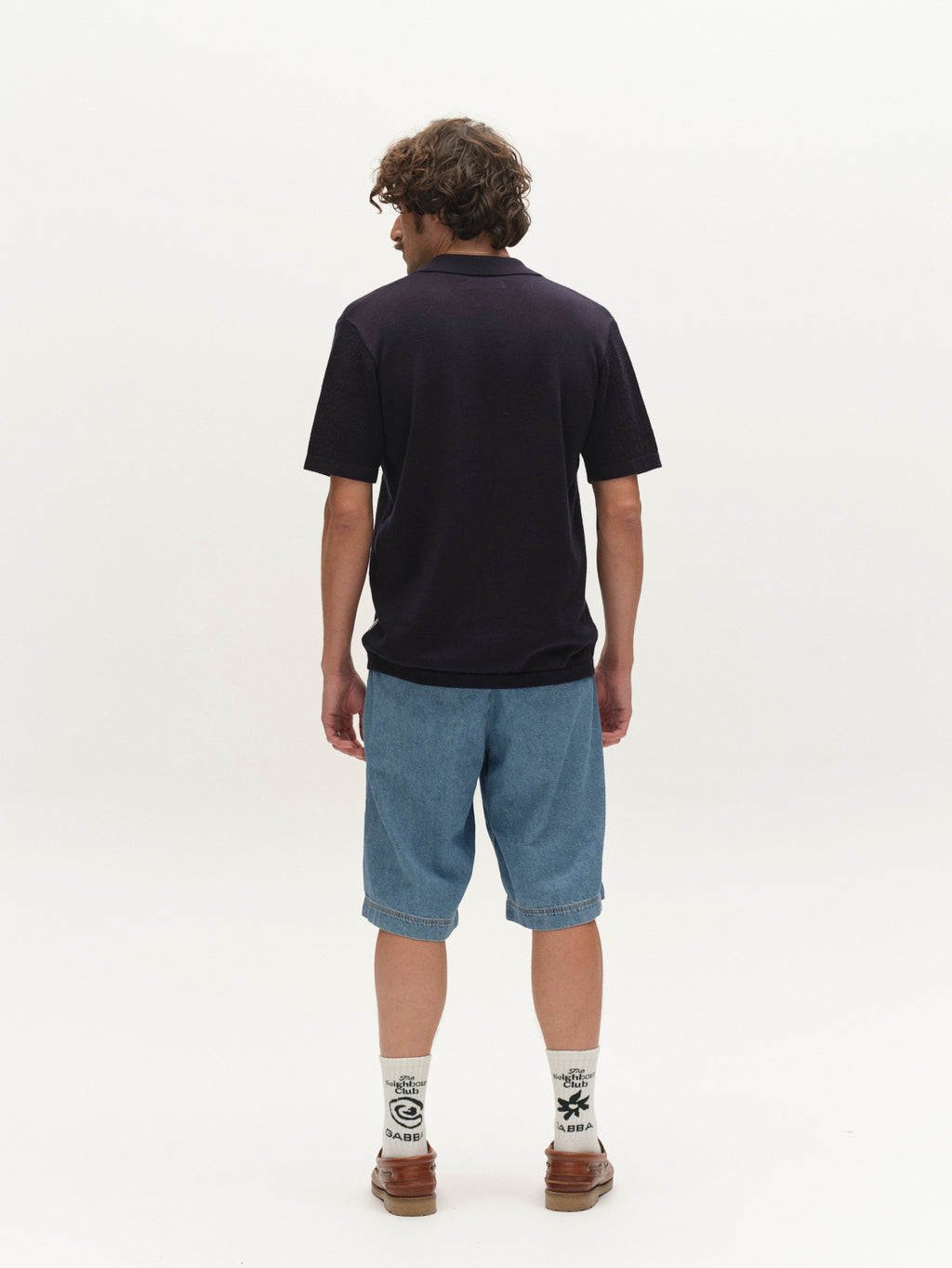 A person with curly hair faces away, wearing a relaxed fit black polo, Gabba's GABmoro PasBrook Shorts in denim wash, white socks with black designs, and brown shoes against a plain white background—a true summer essential look.
