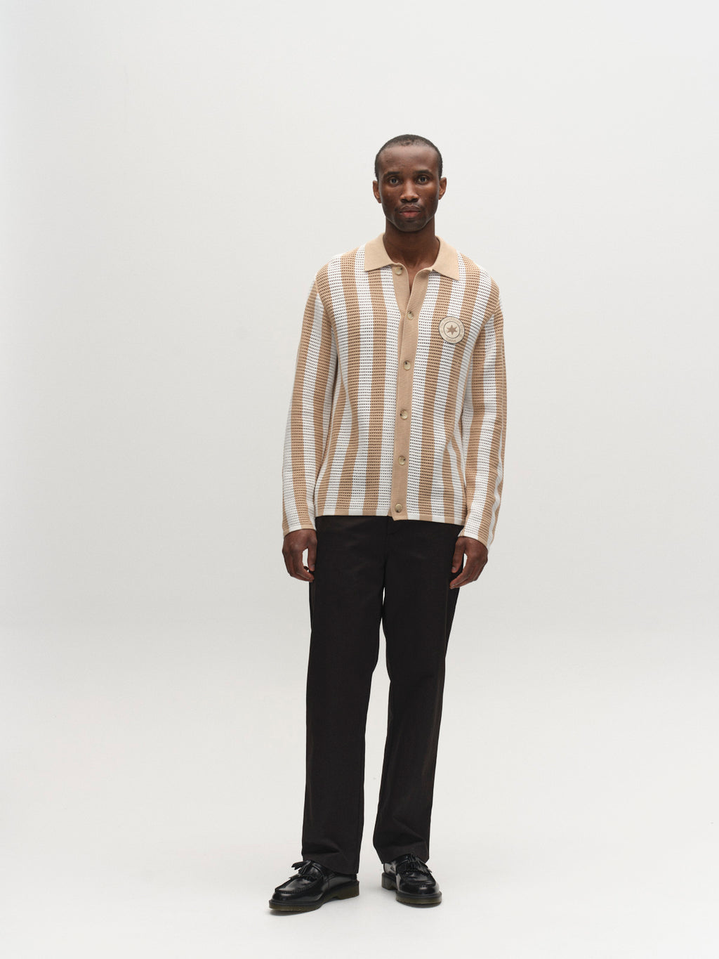 A man stands in a neutral studio, wearing the Gabba GABdezz Bold LS Knit - Beige Stripe cardigan, paired with black pants and shoes. He faces forward with his arms relaxed at his sides against a plain background.