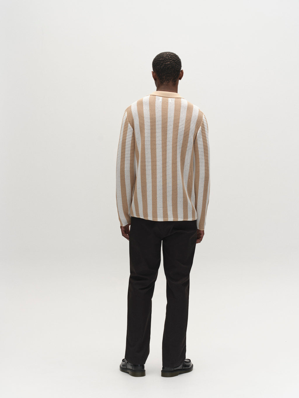 A person stands facing away, wearing the Gabba GABdezz Bold LS Knit in beige stripe, made from 100% cotton, paired with black pants and shoes, against a plain white background.