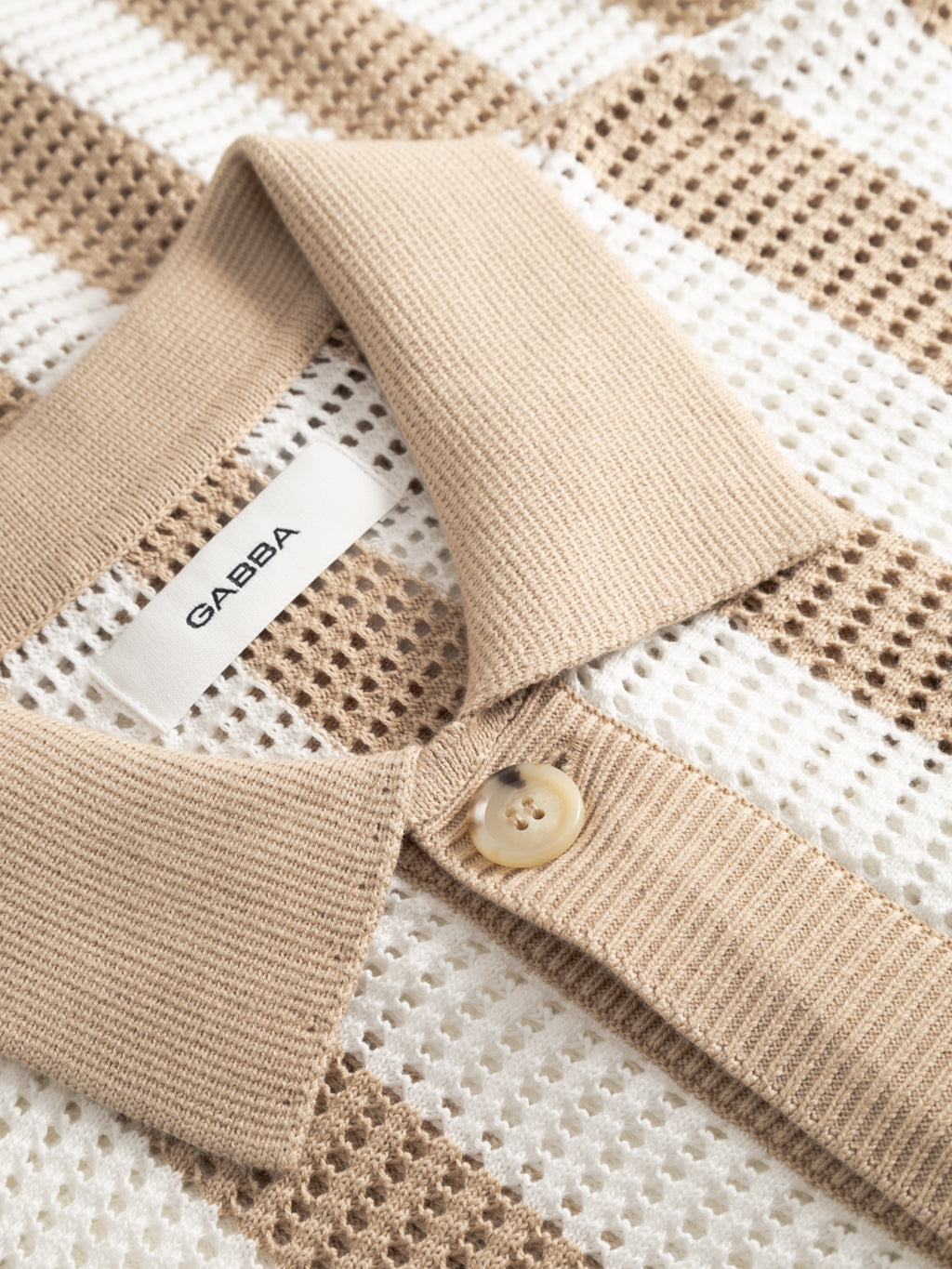 Close-up of the Gabba GABdezz Bold LS Knit in beige and white stripes, featuring a ribbed collar, single button, Gabba label, and mesh-like textured fabric with alternating beige and white stripes.