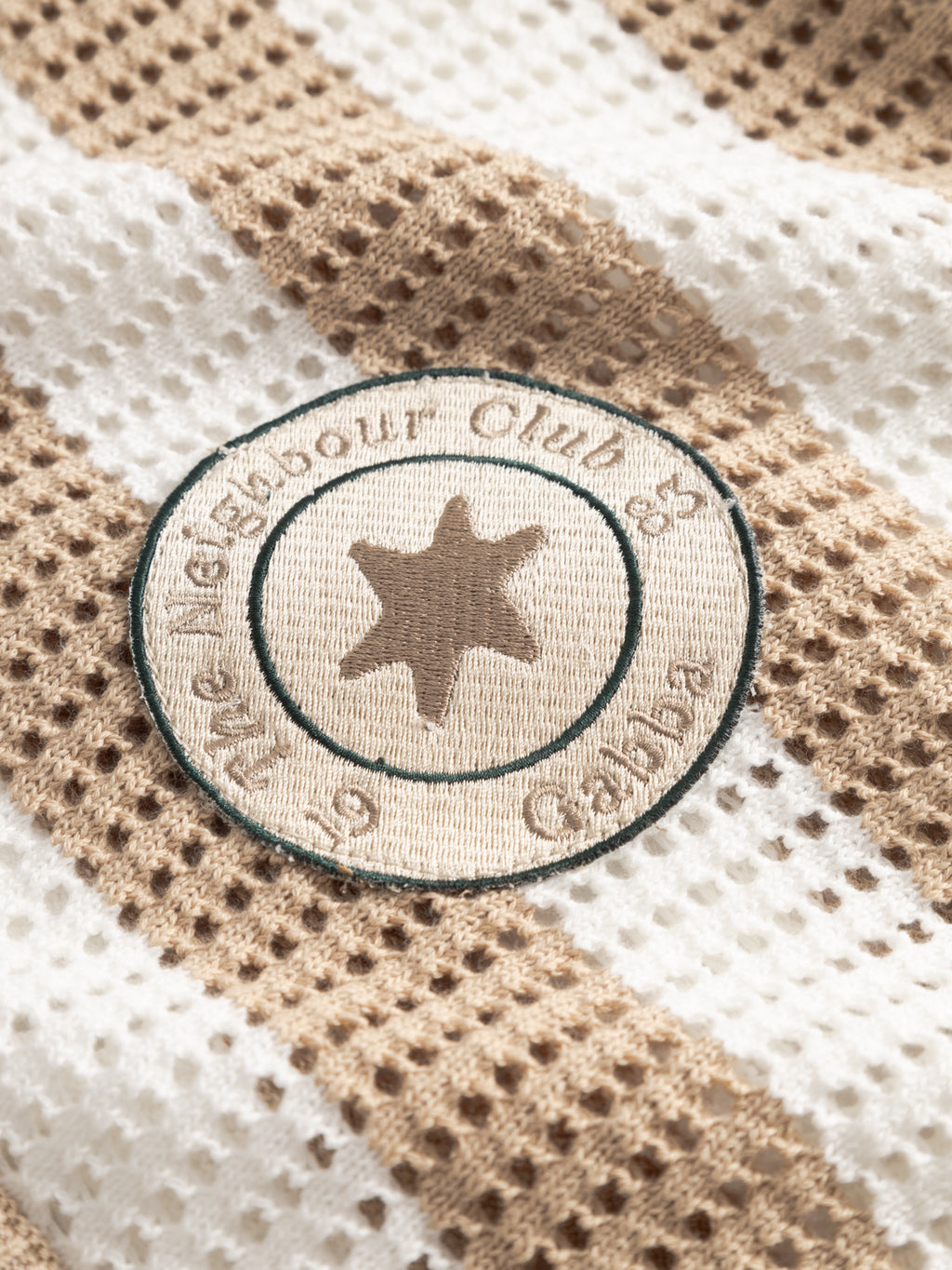 A close-up of the Gabba GABdezz Bold LS Knit in Beige Stripe, showcasing its textured beige and white waffle weave 100% cotton fabric, detailed with a circular embroidered patch featuring a central star and cursive text.