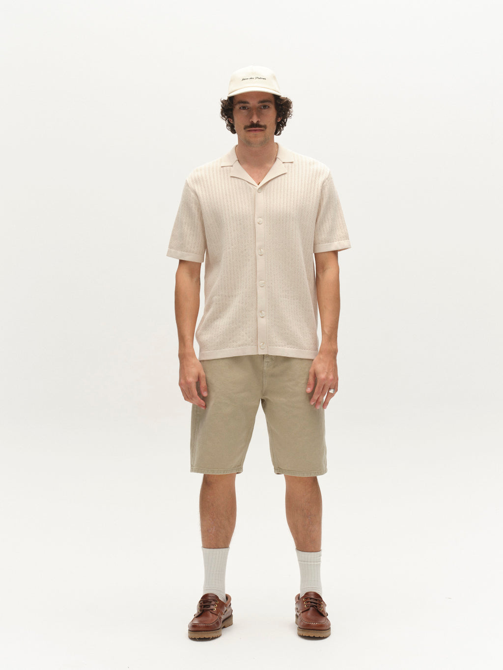 A man with a mustache and curly hair stands against a white background, wearing the Gabba GABgeno Resort Bride SS Knit in Birch, paired with light khaki shorts, white socks, brown shoes, and a beige baseball cap.