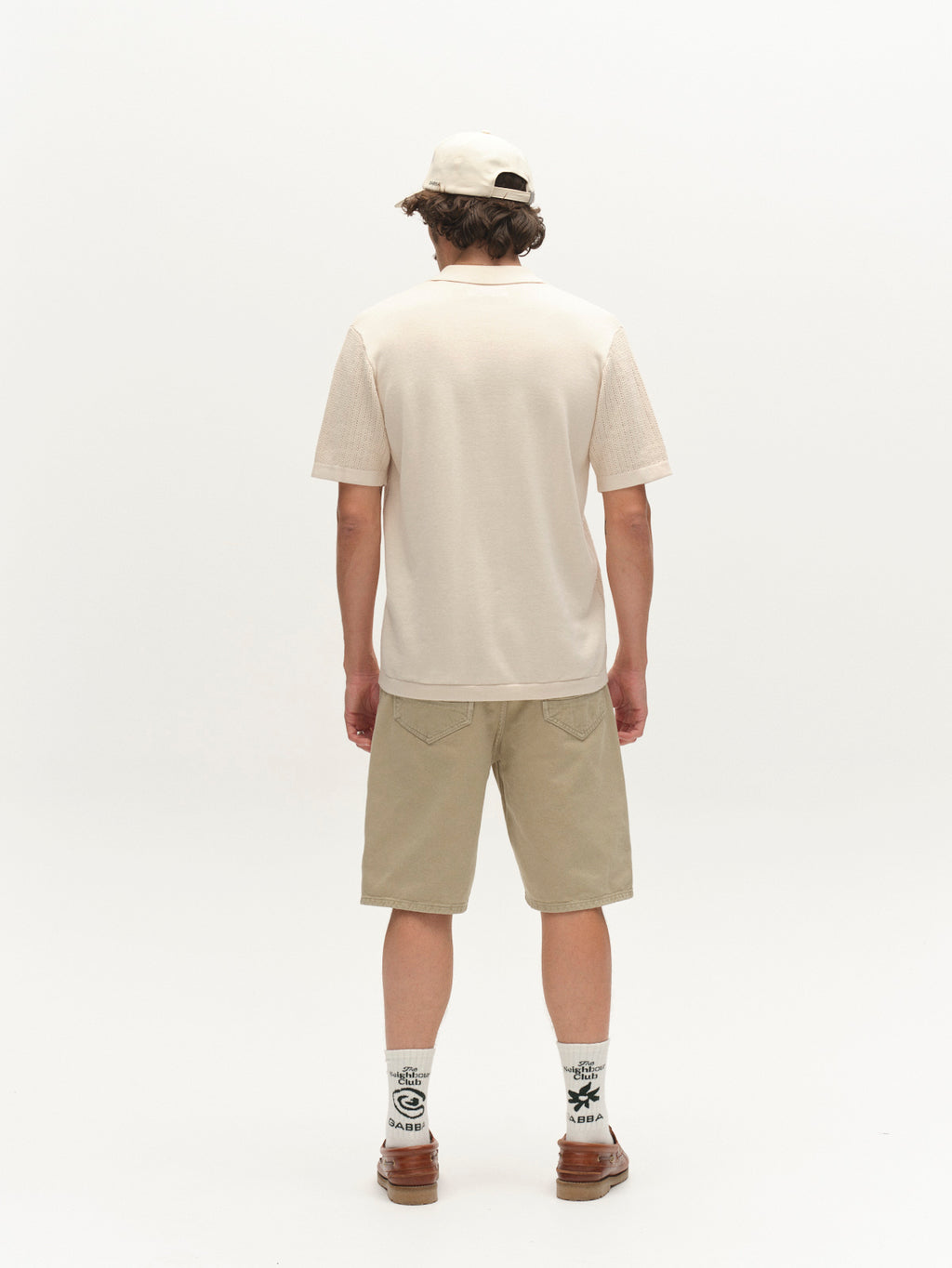A person stands facing away, wearing the Gabba GABgeno Resort Bride SS Knit in Birch, a beige cap, khaki shorts, patterned socks, and brown shoes against a plain white background.