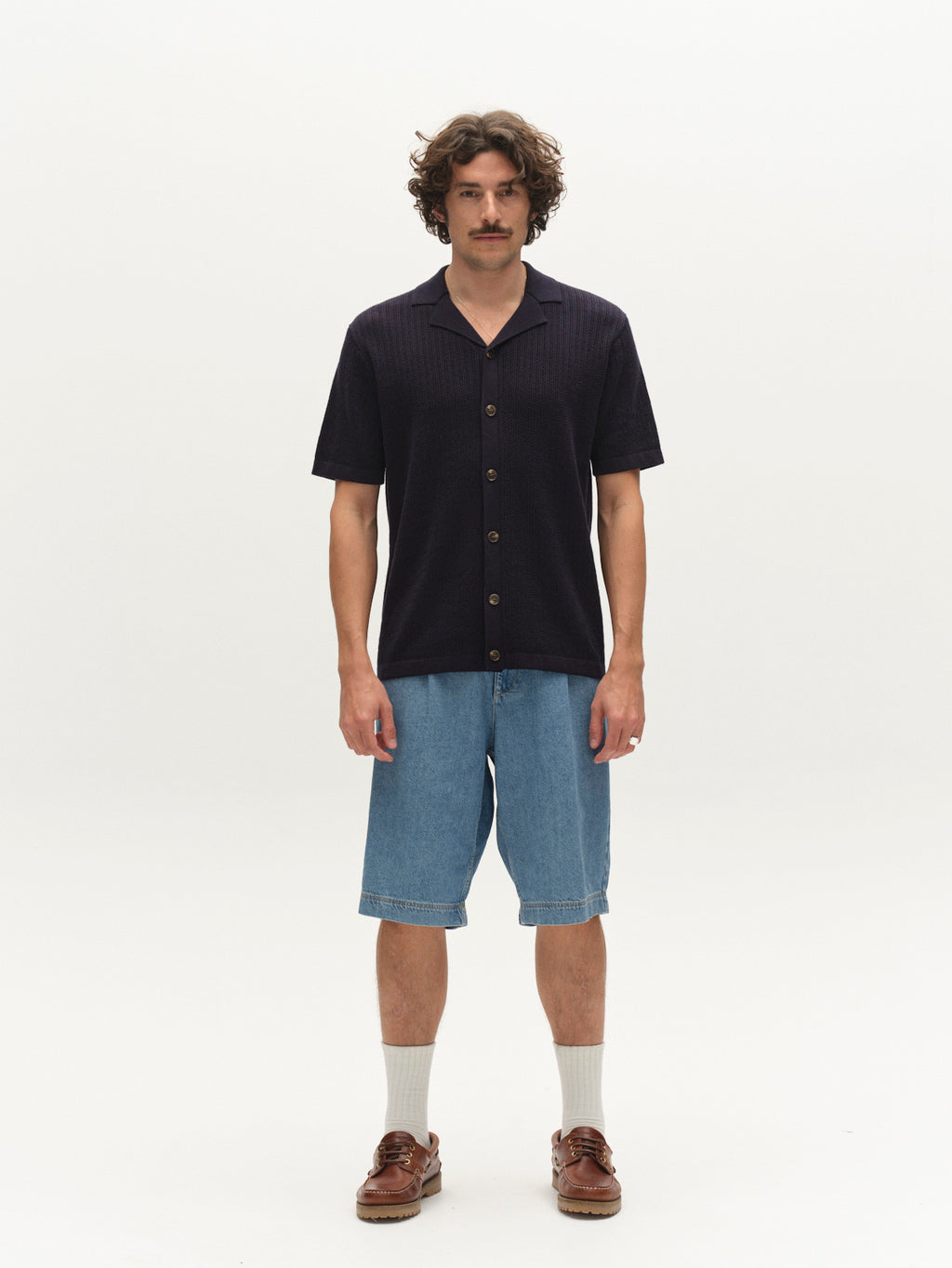 A man with wavy brown hair and a mustache stands facing forward in a Gabba GABgeno Resort Bride SS Knit - Navy shirt, knee-length blue denim shorts, white socks, and brown loafers against a plain white background.