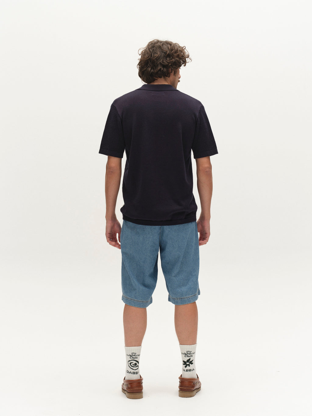 A person with curly hair faces away, wearing the Gabba GABgeno Resort Bride SS Knit in navy, blue knee-length shorts, patterned white socks, and brown shoes against a plain white background.