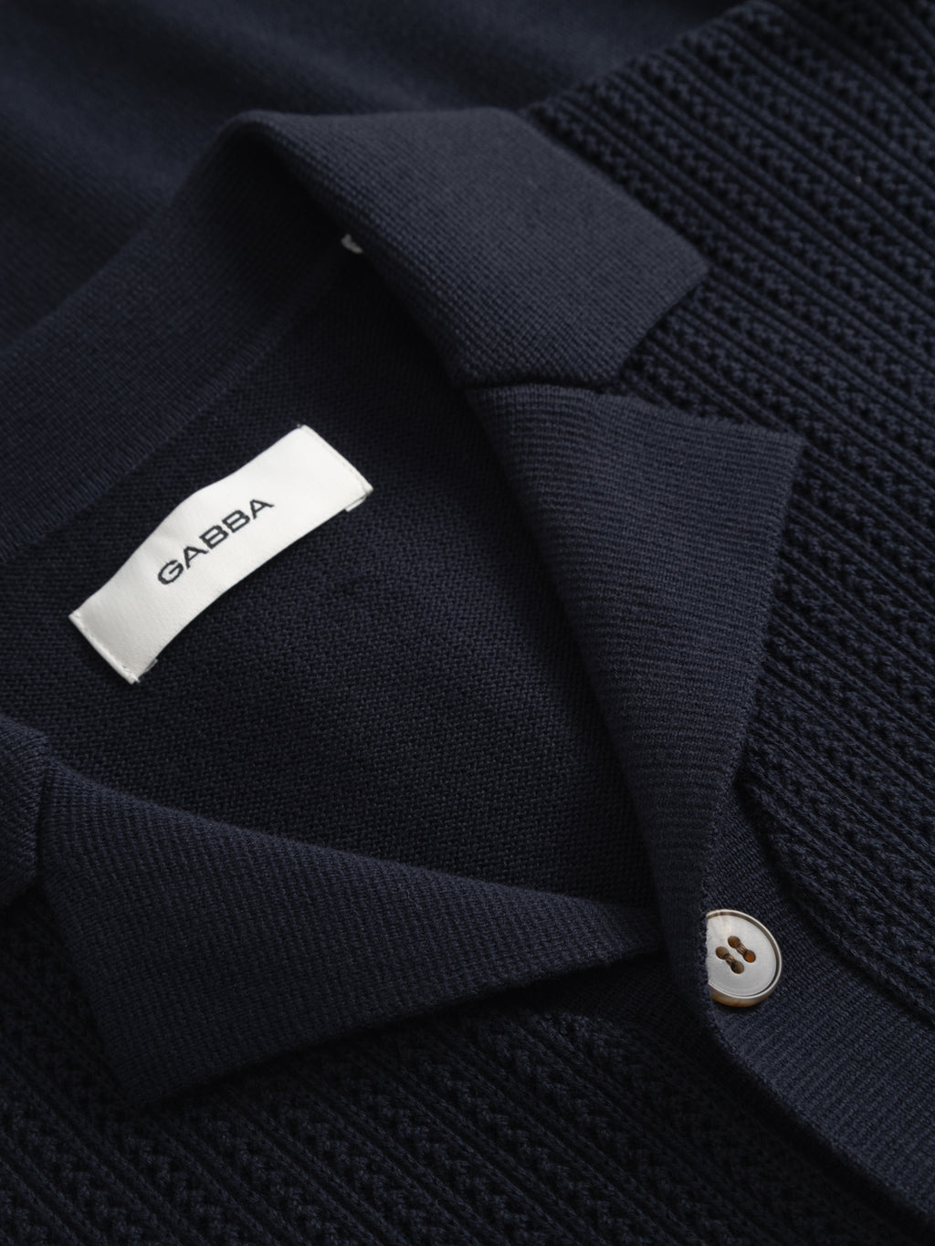 Close-up of the Gabba GABgeno Resort Bride SS Knit shirt in navy, featuring textured knit fabric, a collar, a visible tan button, and an inner white label displaying 'Gabba'.