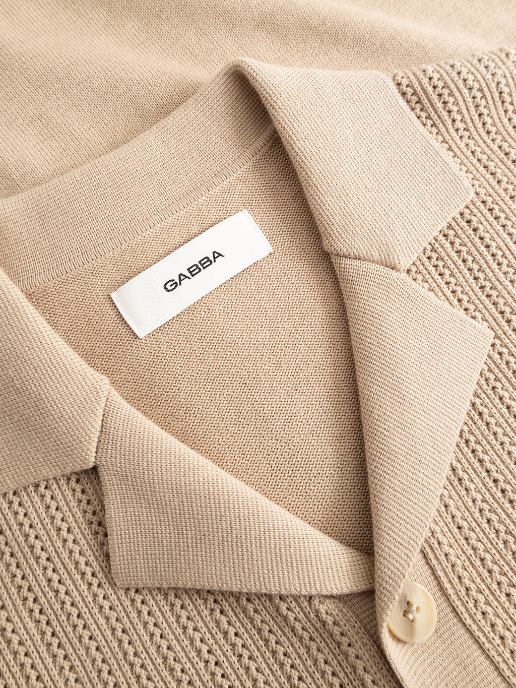 Close-up of the Gabba GABgeno Resort Bride SS Knit in Sand—a beige knit collared shirt with textured pattern, visible buttons, and a white GAËBA label inside the collar.
