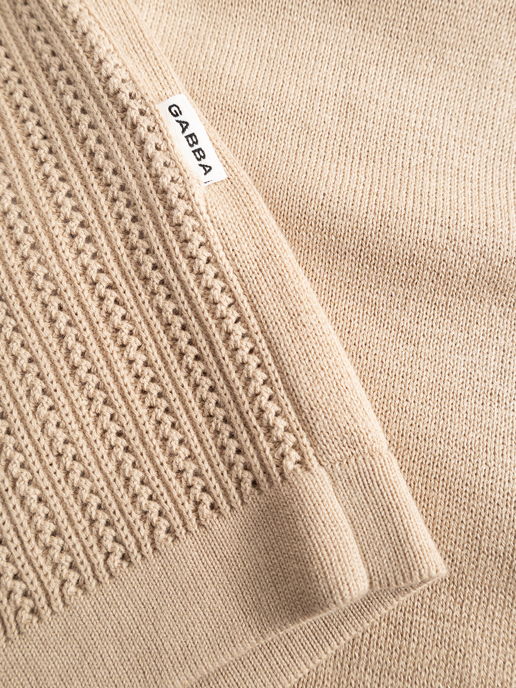 Close-up of the GABgeno Resort Bride SS Knit in Sand by Gabba, showing beige fabric with ribbed and smooth textures. A white “GABBA” label is sewn onto the ribbed section.