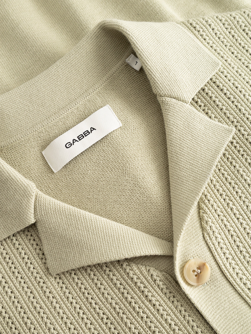 A close-up of the Gabba GABgeno Resort Bride SS Knit in light green shows the short-sleeve cotton knit texture, stitching details, button, and white Gabba label.