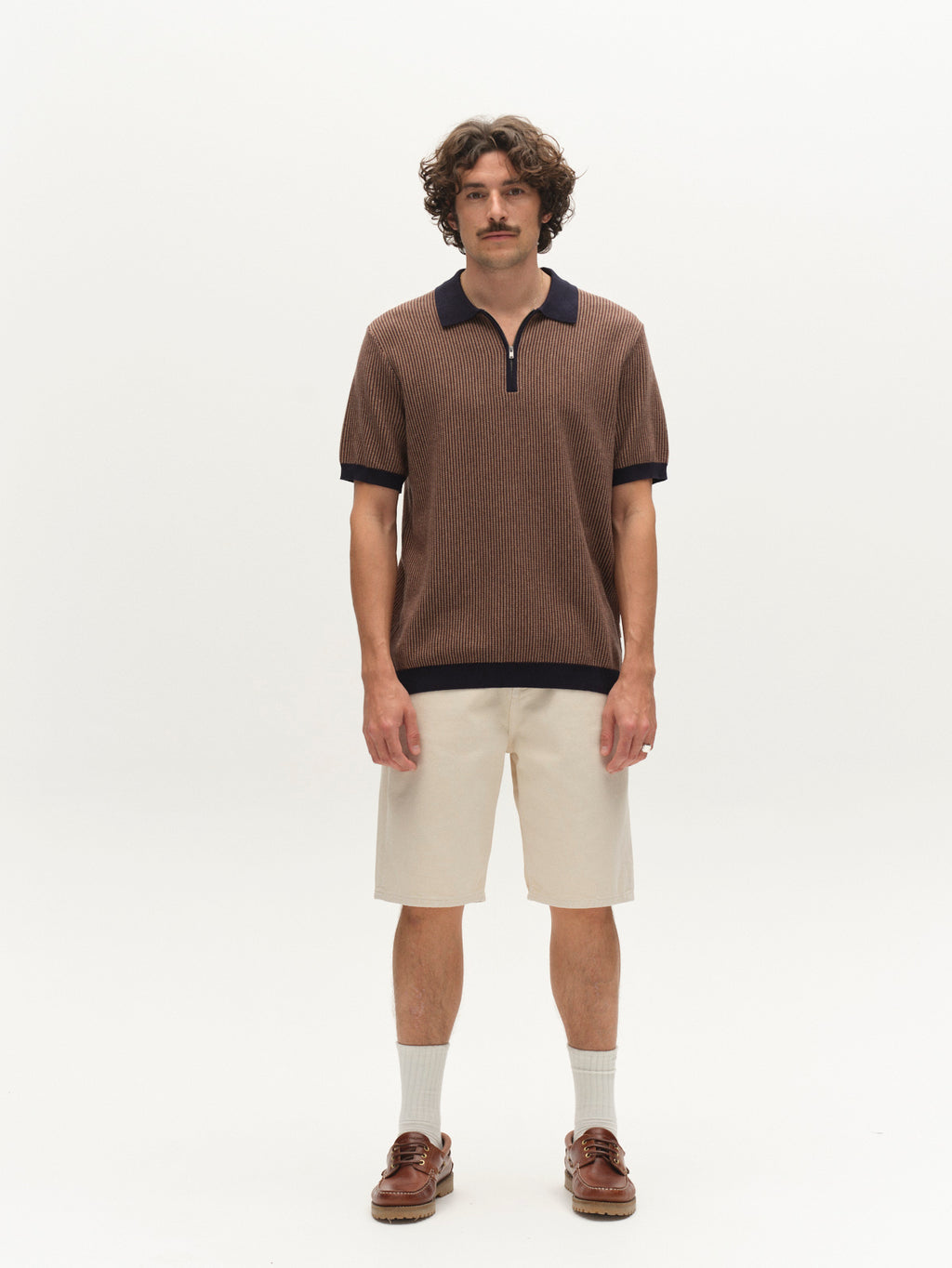 A man with curly hair and a mustache stands against a white background, wearing the Gabba GABoswald Retro SS Knit polo in navy, beige shorts, light socks, and brown boat shoes.