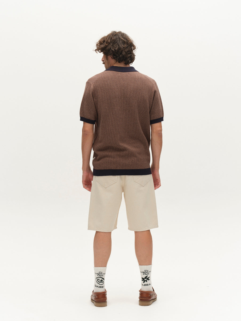 A person with curly hair stands facing away, wearing the Gabba GABoswald Retro SS Knit in Navy paired with cream knee-length shorts, patterned white socks, and brown shoes against a plain white background.