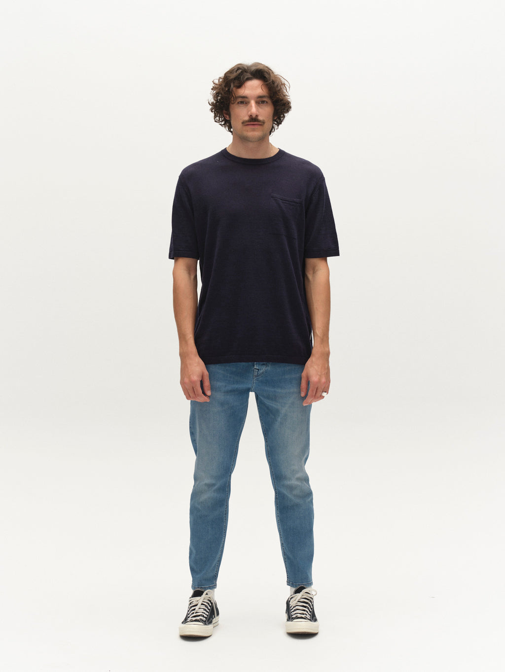 A man with curly hair and a mustache faces forward, wearing Gabba's GABvento Dush SS Knits in navy, paired with light blue jeans and black and white sneakers, standing against a plain white background.