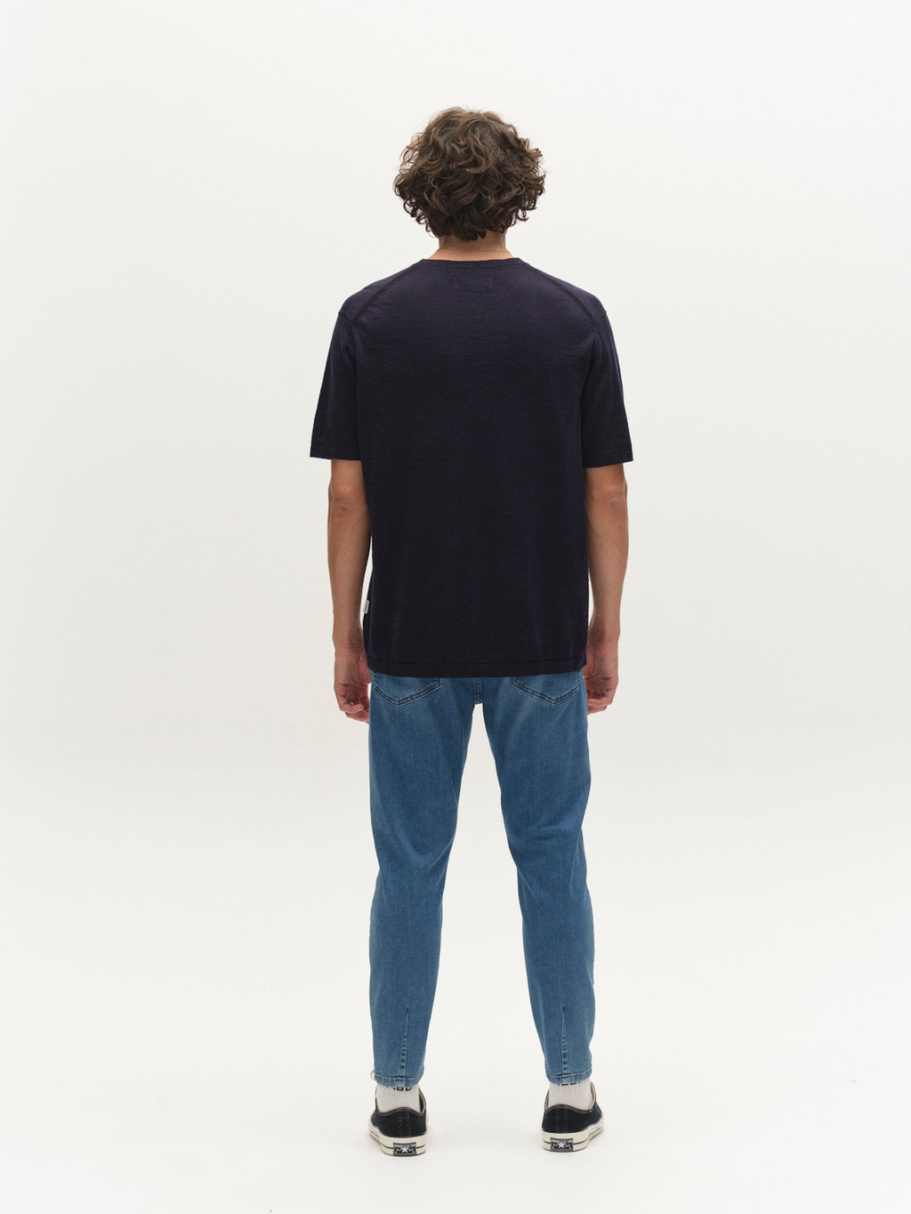 A person with curly hair stands facing away, wearing the Gabba GABvento Dush SS Knits in navy, paired with blue jeans and black-and-white sneakers against a plain white background.