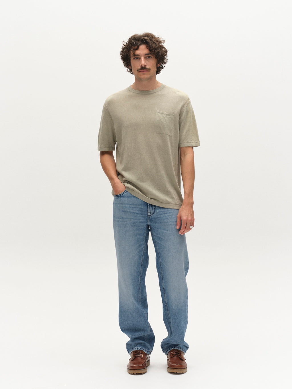 A man with curly hair and a mustache faces forward, wearing the Gabba GABvento Dush SS Knits in light green, light blue jeans, and brown shoes against a plain white background.