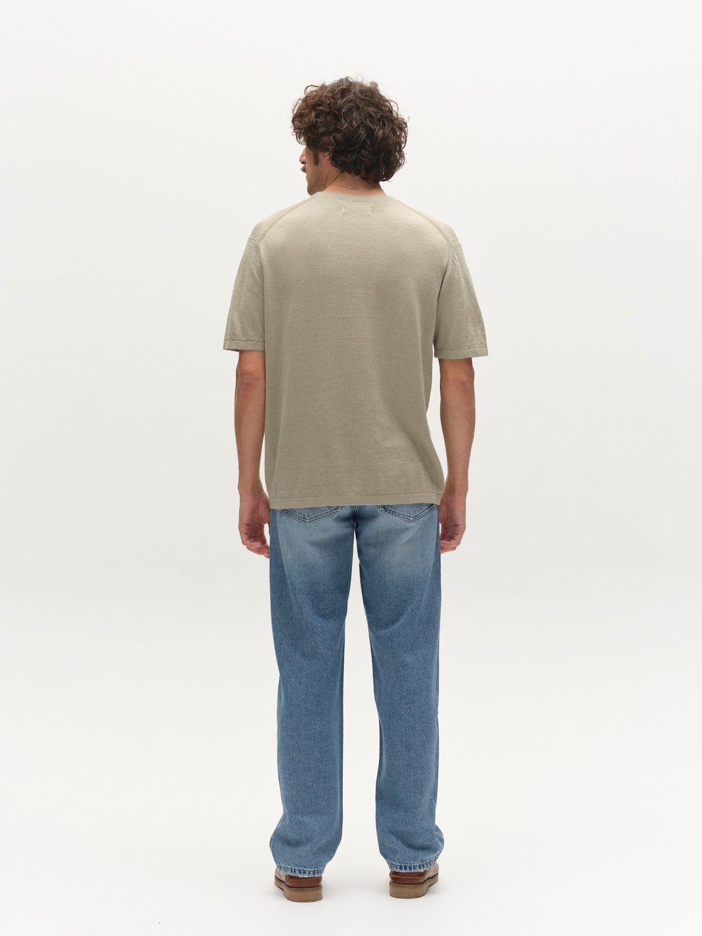A man with curly hair faces away, wearing the Gabba GABvento Dush SS Knits in light green, paired with light blue jeans and brown shoes against a plain white background.
