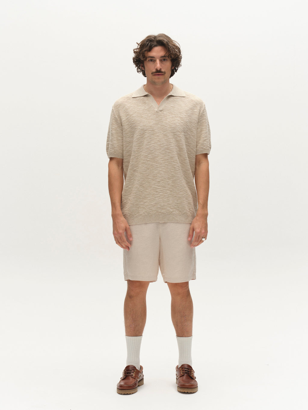 A man with wavy hair and a mustache stands against a white background, wearing the Gabba GABcross Alto SS Knit in Ecru, light beige shorts, white socks, and brown boat shoes.