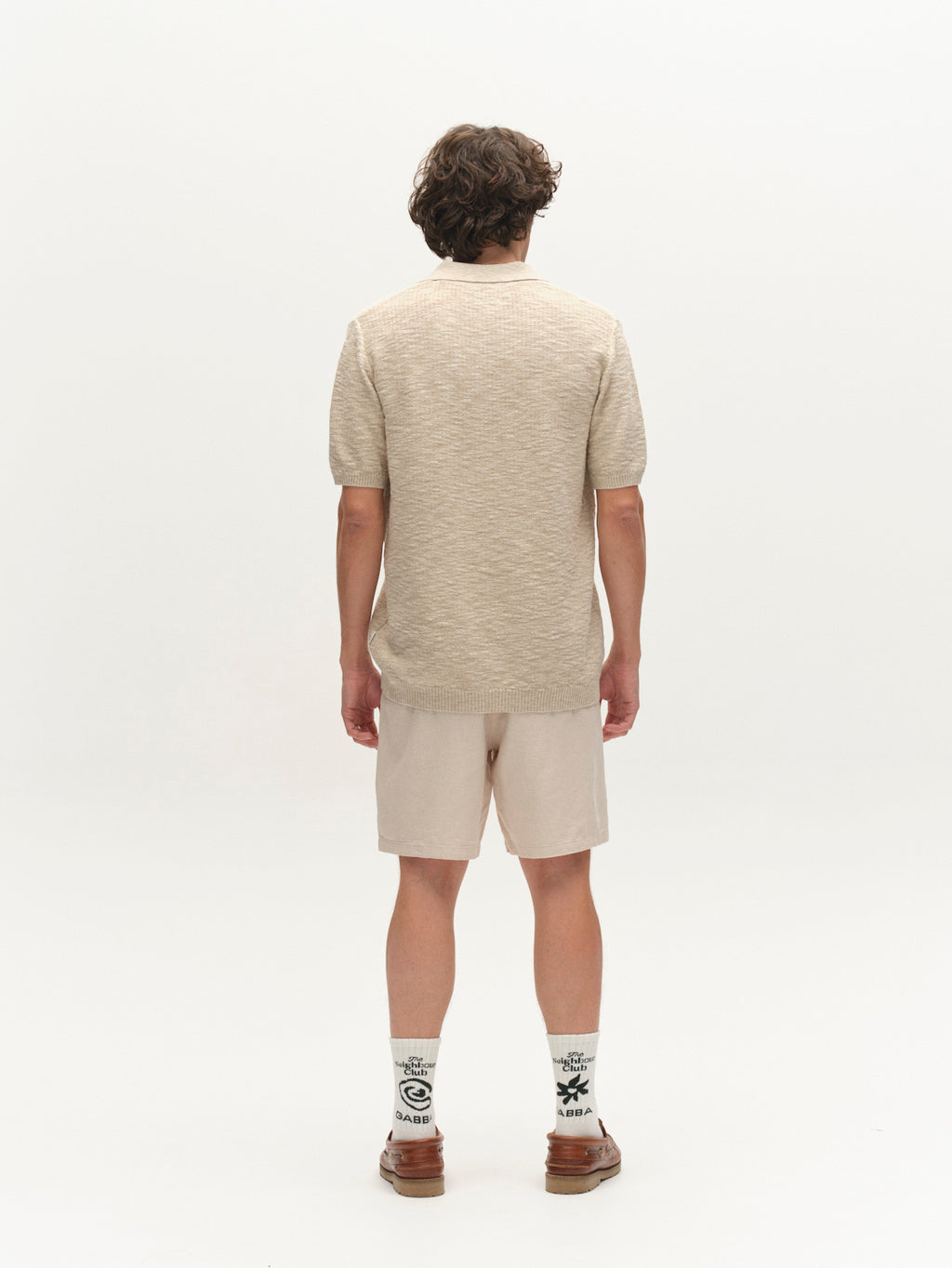 A person with wavy hair stands facing away, wearing the Gabba GABcross Alto SS Knit in ecru, light beige shorts, patterned white socks, and brown sandals against a plain white background.