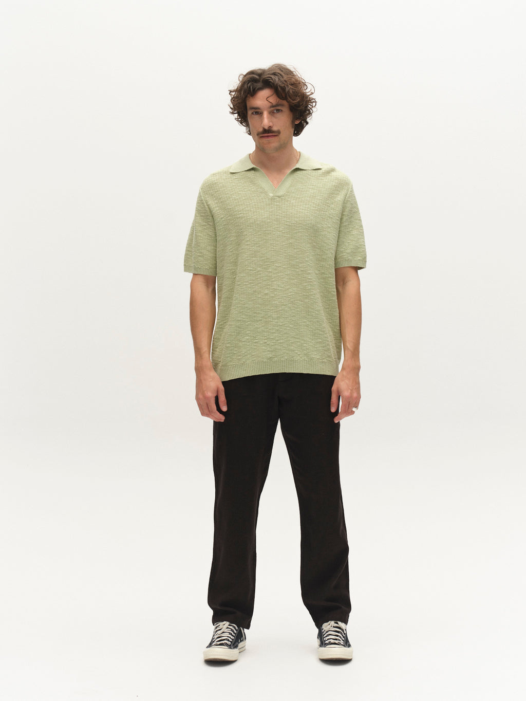 A man with curly hair and a mustache stands against a plain white background, wearing the Gabba GABcross Alto SS Knit in Alfa Green, dark pants, and black sneakers with white soles.