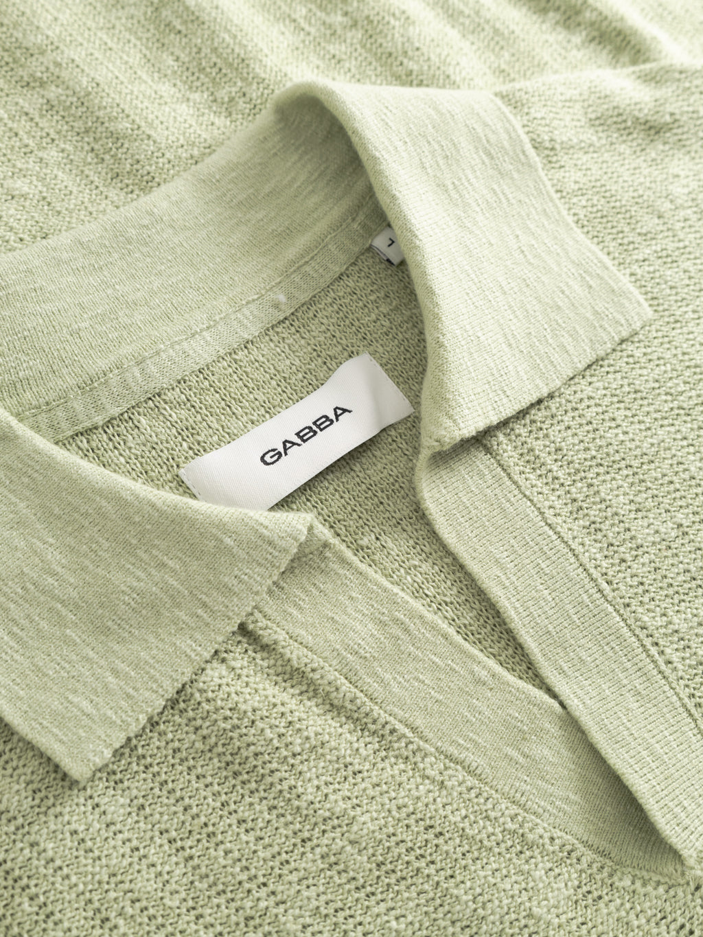Close-up of a light green textured collared shirt, the GABcross Alto SS Knit in Alfa Green by Gabba, neatly folded with a white brand label visible.
