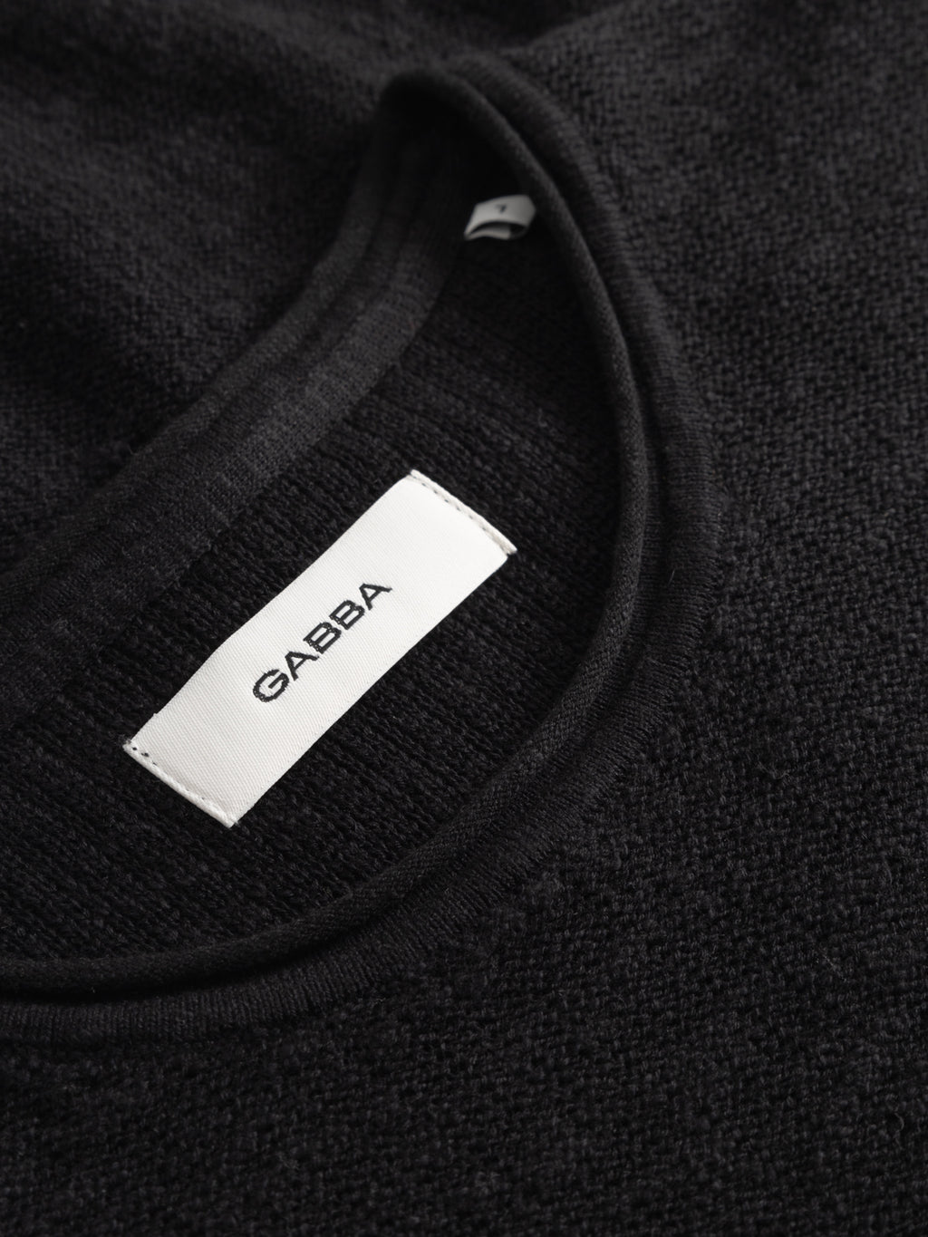 Close-up of a black textured GABnigel Alto SS Knit by Gabba, featuring a white fabric label sewn inside the neckline with the brand name Gabba in black capital letters.
