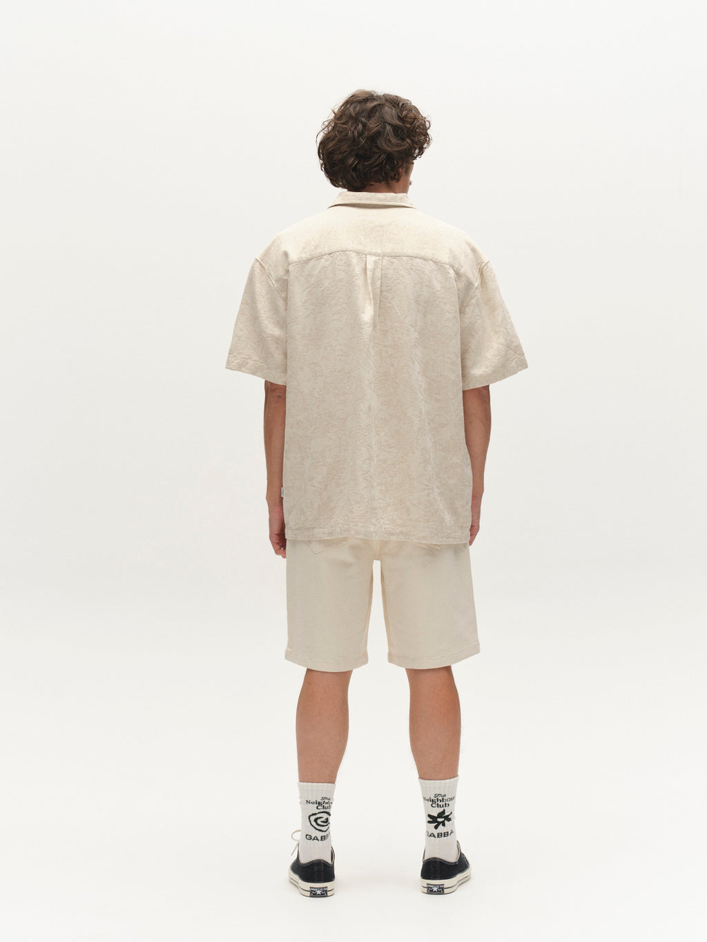 A person with curly hair stands facing away, wearing a short-sleeve beige shirt and Gabba GABmox Twill Shorts in ecru, paired with white socks featuring black graphics and black sneakers, set against a plain white background.
