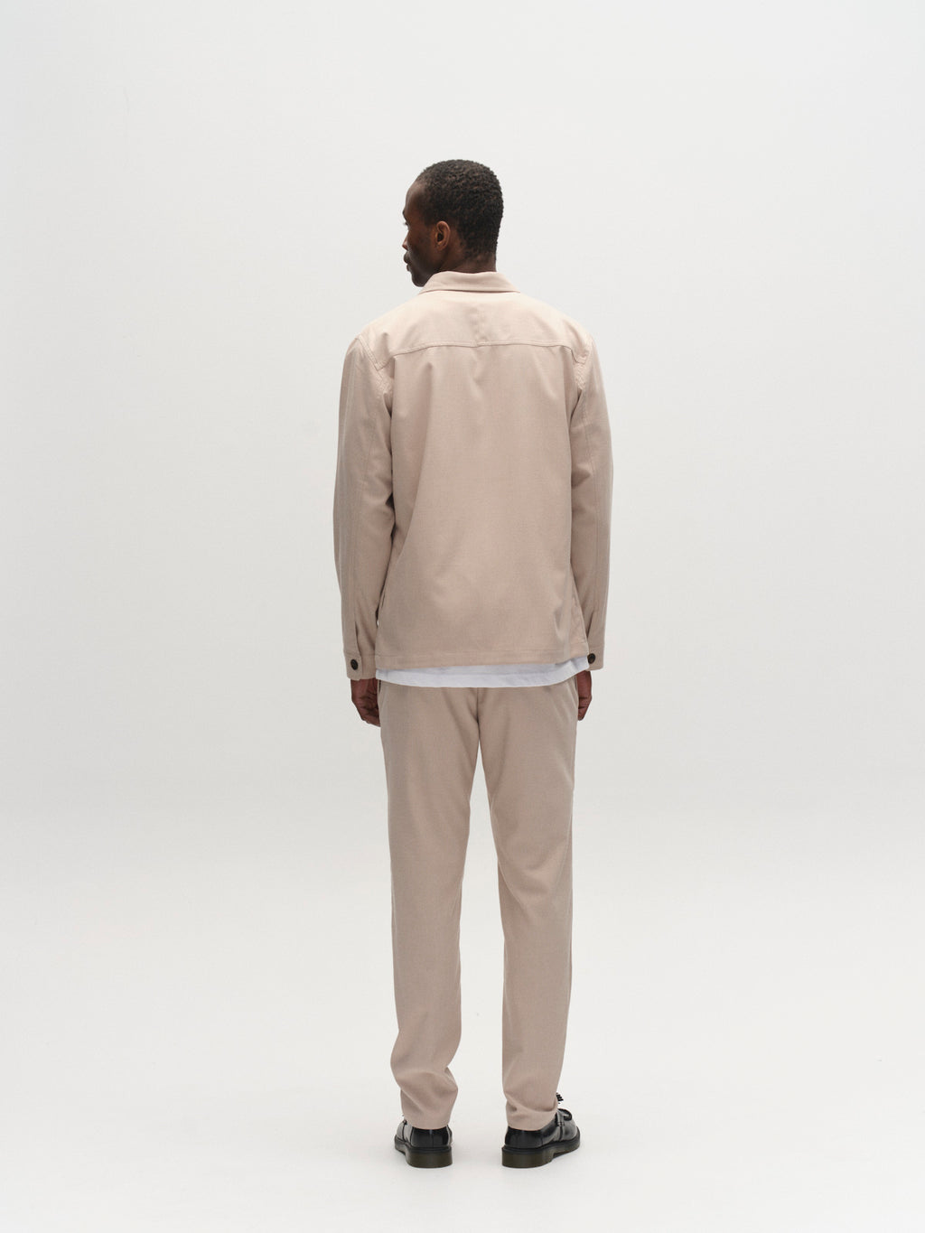 A man faces away from the camera against a plain white background, wearing Gabba’s GABpaul Deme Pant in Birch with a matching jacket, a white shirt, and black sandals.