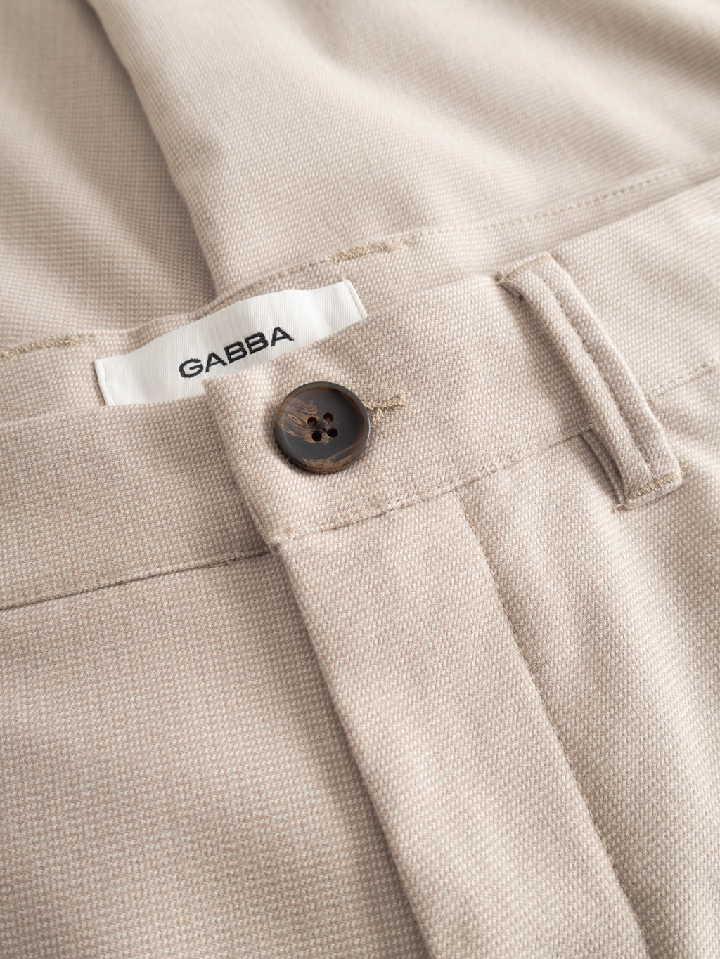 Close-up of Gabba GABpaul Deme Pant - Birch: beige slim-fit trousers with brown button, belt loop, and white Gabba label, crafted from micro-pattern woven fabric for texture.