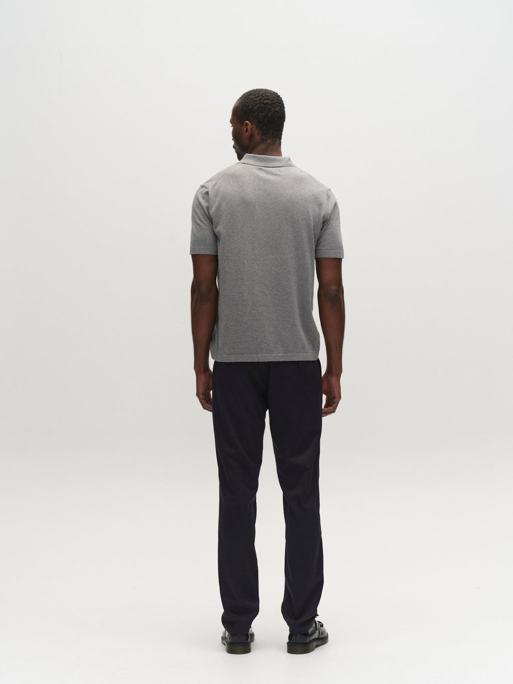 A man faces away from the camera, wearing a gray two-tone yarn short-sleeve polo, Gabba GABpaul Deme Pant in dark navy, and black shoes against a plain white background.
