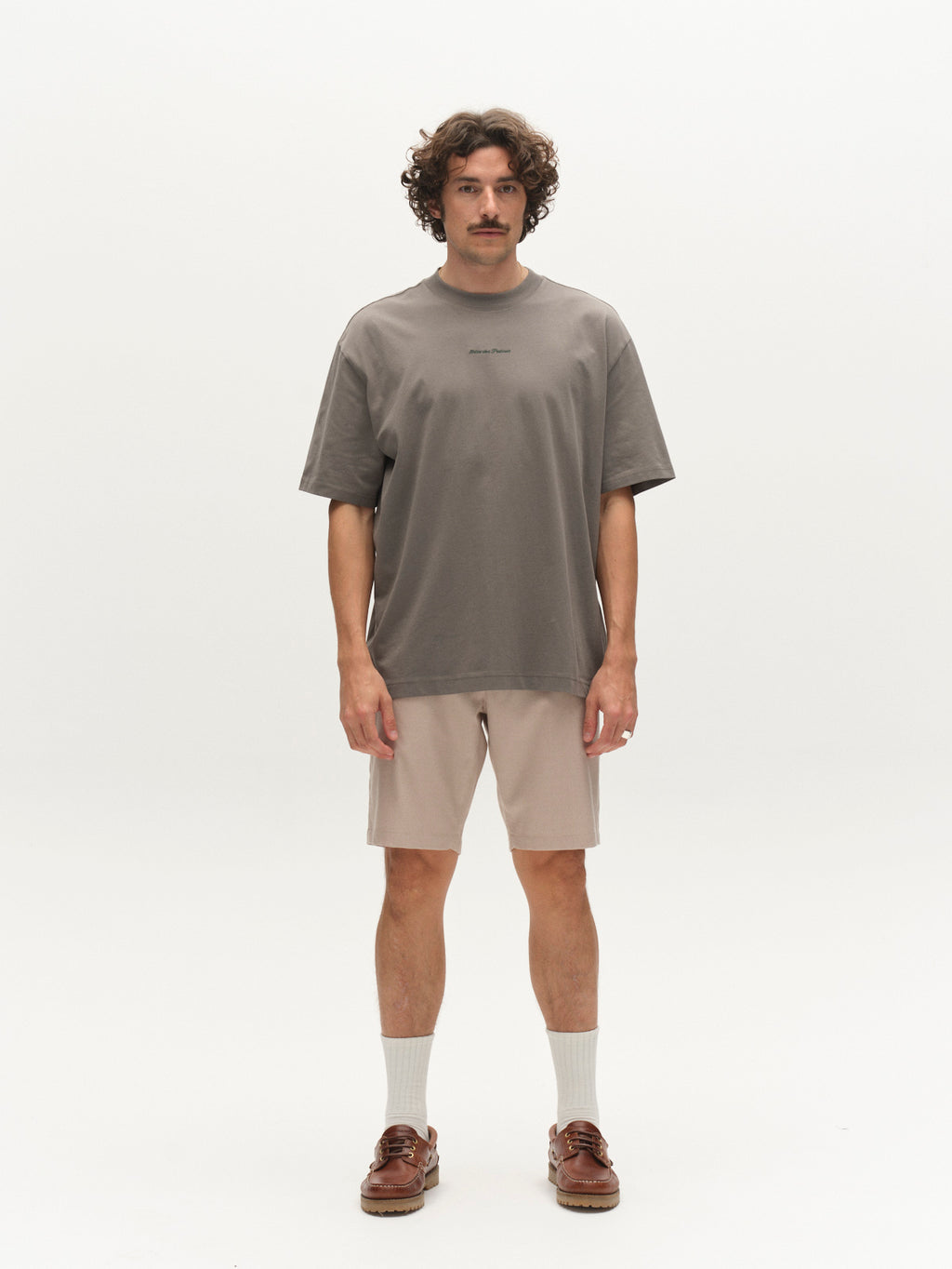 A man with curly hair and a mustache stands against a plain white background, wearing a loose gray t-shirt, Gabba GABjet Deme Shorts - Birch, white socks, and brown boat shoes. He faces forward with a neutral expression.