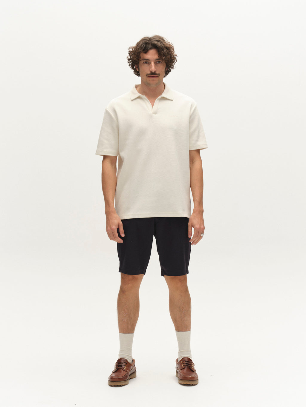 A man with curly hair and a mustache stands against a white background, wearing a cream short-sleeve collared shirt, Gabba GABjet Deme Shorts in Dark Navy, light socks, and brown boat shoes.