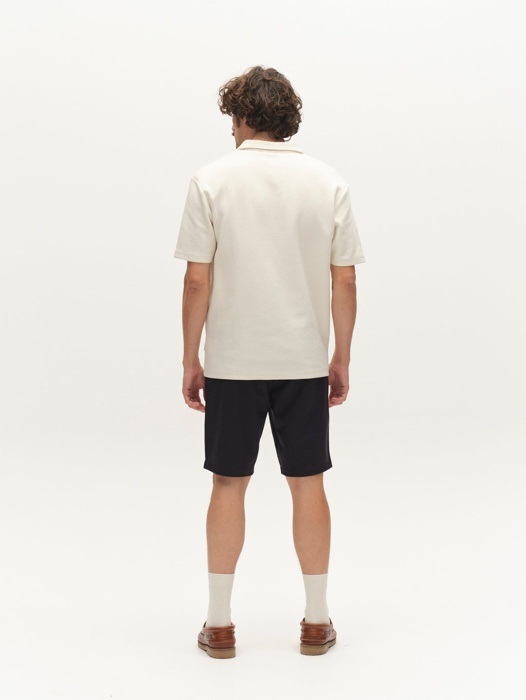 A man with curly brown hair, facing away, wears a cream polo shirt, white socks, brown loafers, and Gabba GABjet Deme Shorts in Dark Navy against a plain white background.
