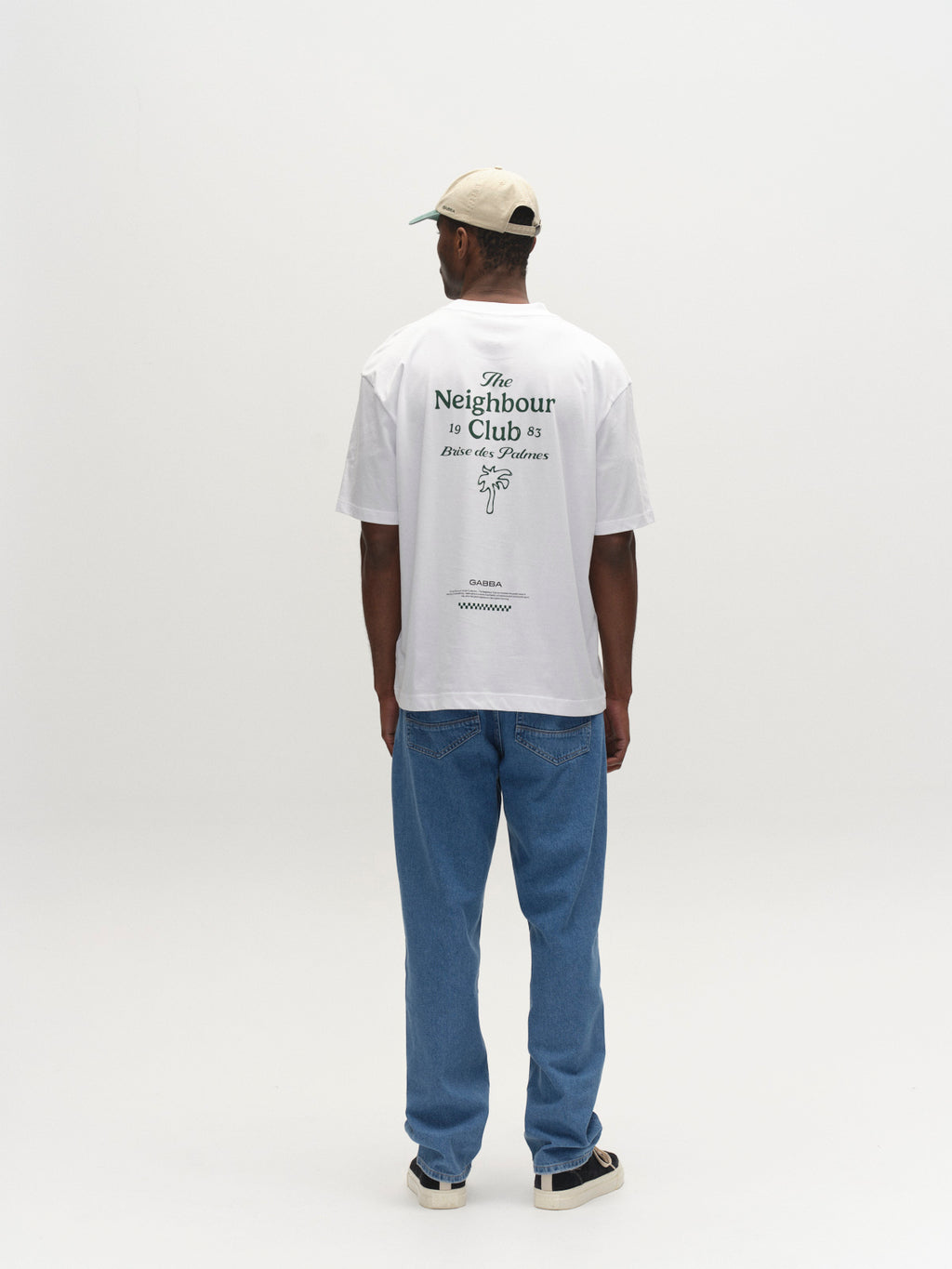 A man faces away, wearing the Gabba GABnigel NBH SS T-Shirt GOTS - White (regular fit, organic cotton, printed text), blue jeans, beige sneakers, and a light cap against a plain white background.