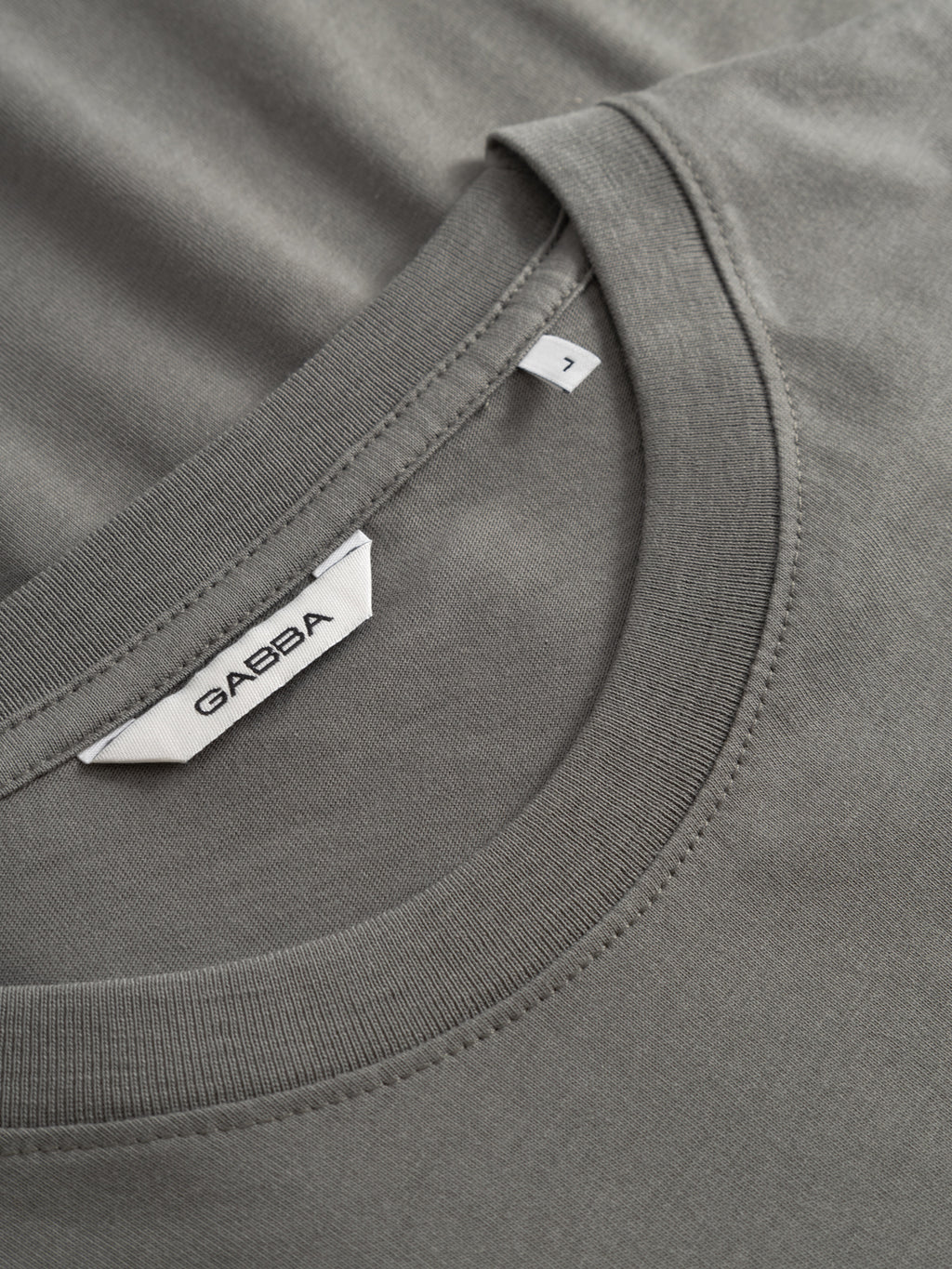 Close-up of the Gabba GABnigel NBH SS T-Shirt GOTS in Castor Gray, featuring a white Gabba label and size 7 tag inside the collar; soft single jersey fabric and stitching are clearly visible.