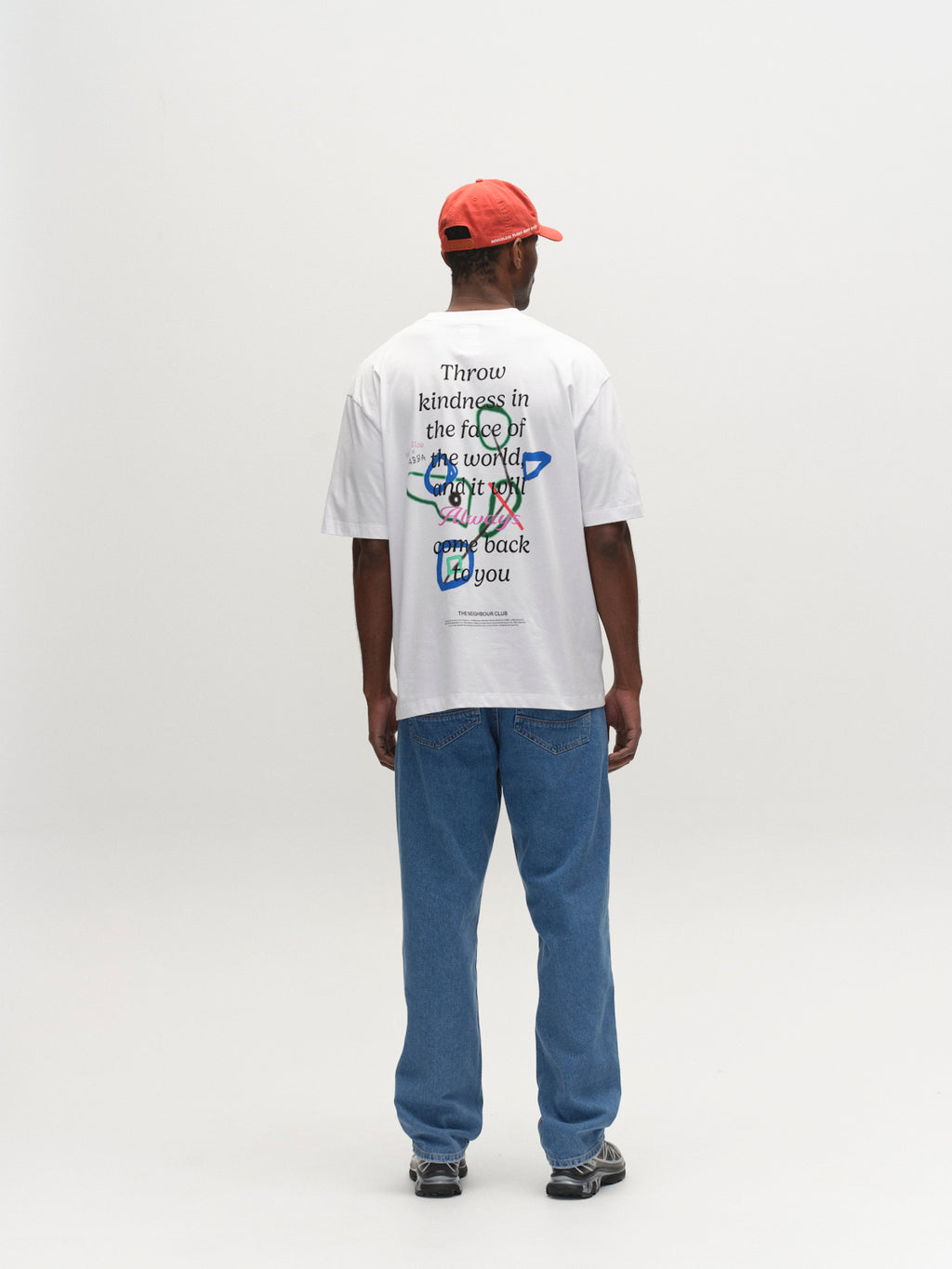 A person wearing a red cap, blue jeans, sneakers, and a Gabba GABnigel Boxy Nando SS T-Shirt GOTS in white stands facing away in a plain, well-lit studio.