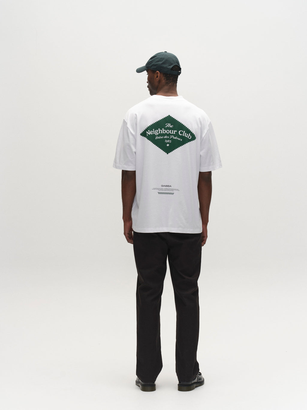 A person faces away in a Gabba GABnigel Boxy NBH SS T-shirt GOTS - White, featuring a green diamond Neighbour Club print, paired with dark pants, a dark green cap, and black shoes in a plain white studio.