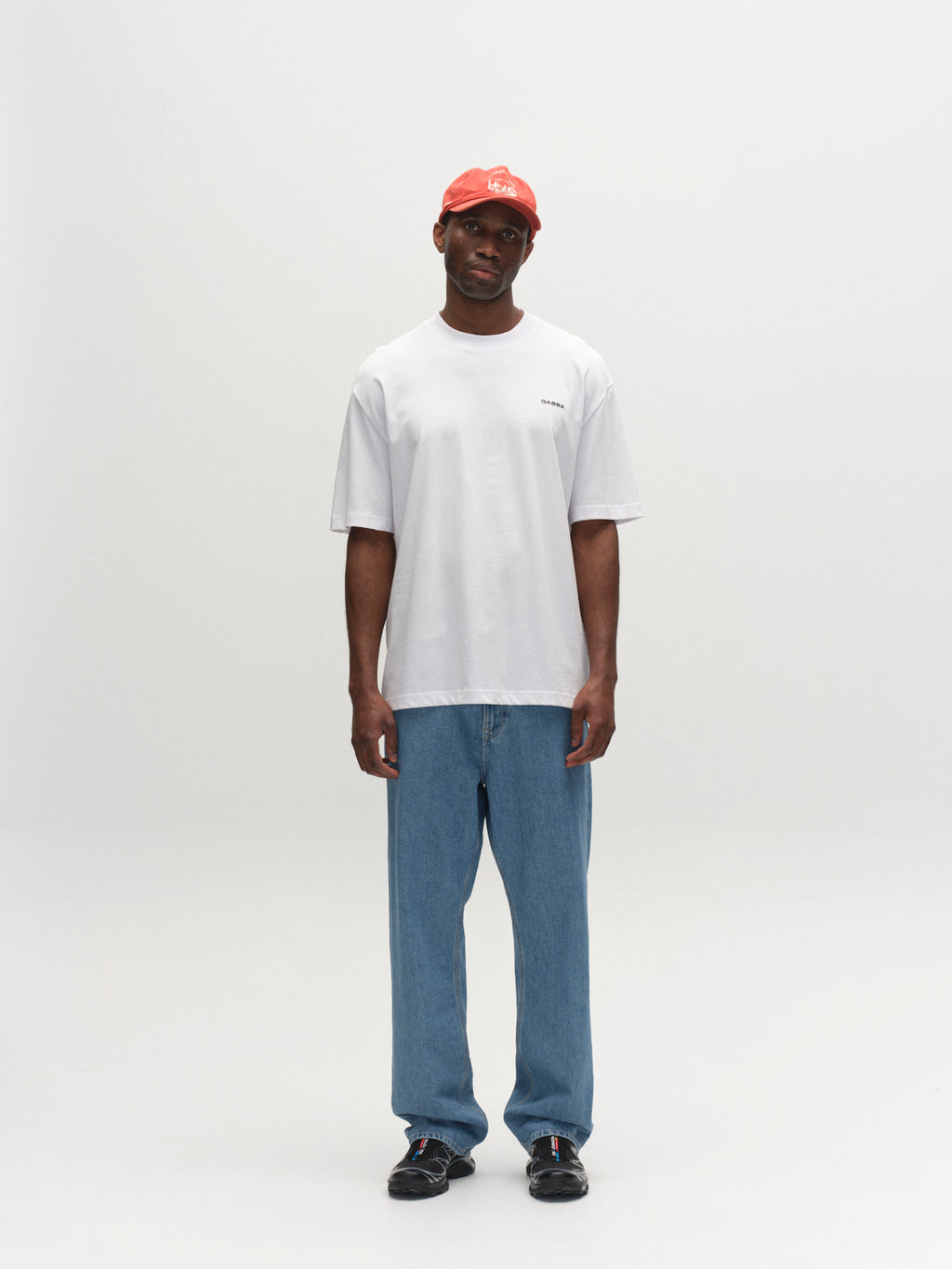 A man wearing a Gabba GABnigel Boxy Logo GOTS SS white t-shirt, blue jeans, black shoes, and a red cap stands against a white background, looking straight ahead with his arms relaxed by his sides.