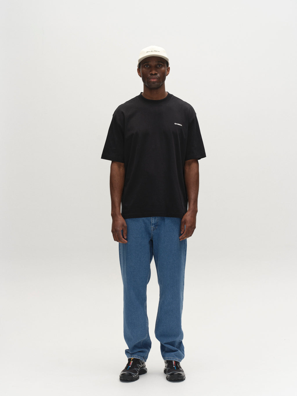 A man stands facing forward against a white background, wearing the Gabba GABnigel Boxy Logo GOTS SS T-shirt in black, paired with blue jeans, black shoes, and a white baseball cap.