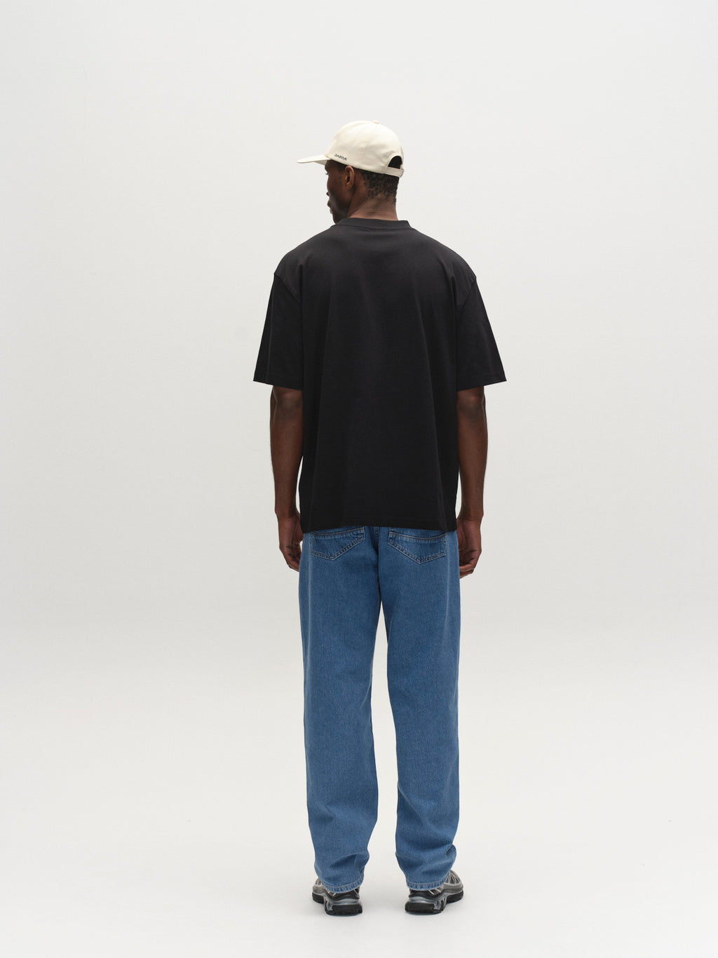 A man faces away from the camera, wearing a Gabba GABnigel Boxy Logo GOTS SS black t-shirt, white cap, blue jeans, and black sneakers against a plain light background.