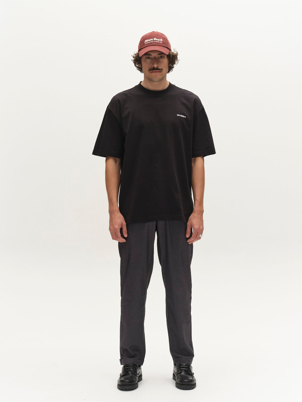 A man with a mustache faces forward in a black t-shirt, Gabba GABjoe Turnup Fin Pant - Lt. Grey chinos, black shoes, and a red Good Good cap against a plain white background.
